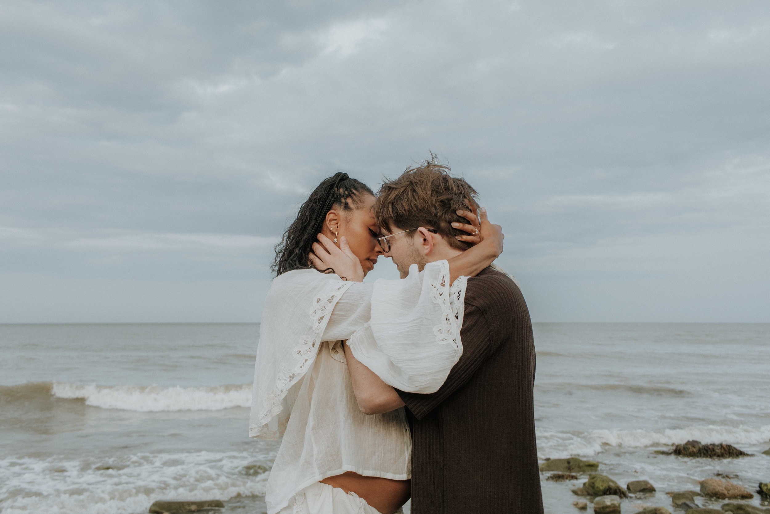 A couple stands close together on a beach, holding each other with foreheads and noses touching, under a cloudy sky.
