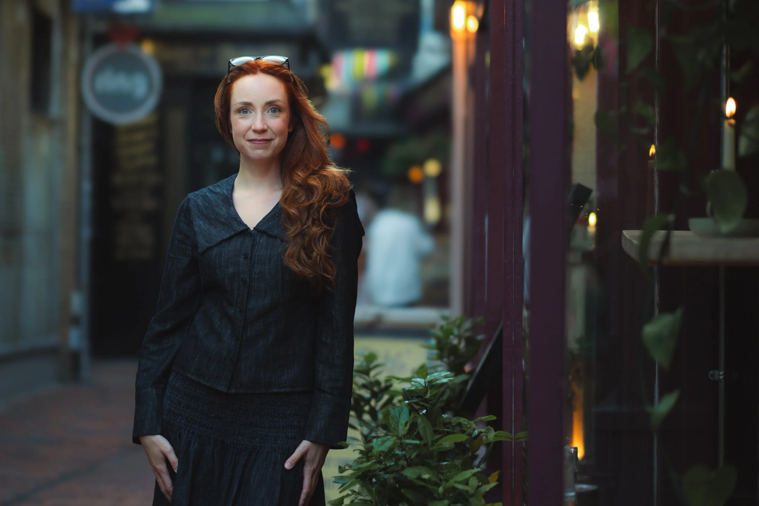 A woman with red hair and glasses resting on her head, wearing a black dress, standing outdoors at dusk near a building with reflections of lights on the window.