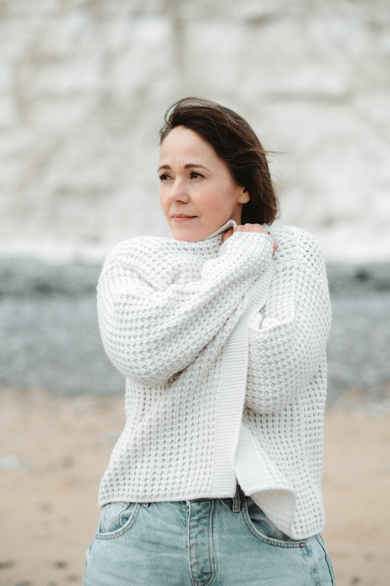 A woman in a white knitted sweater and light blue jeans adjusting her collar outdoors, with a blurred stone wall and beach in the background.