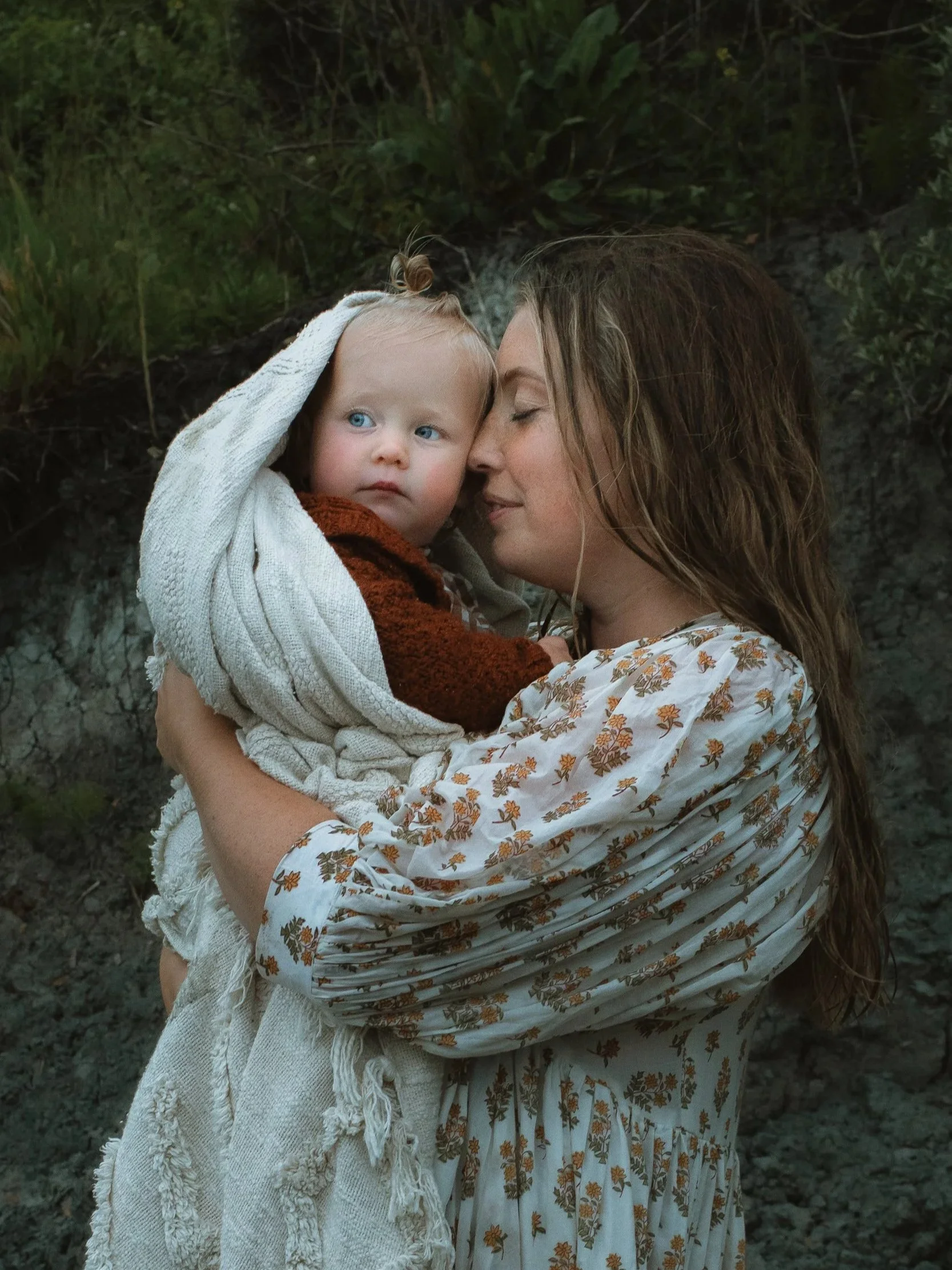 A woman holding a young child wrapped in a blanket outdoors with trees and greenery in the background.