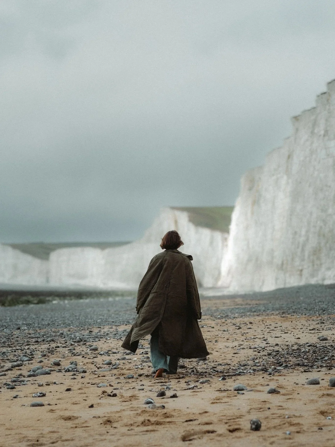 A person walking on a sandy beach with white chalk cliffs in the background under cloudy sky.
