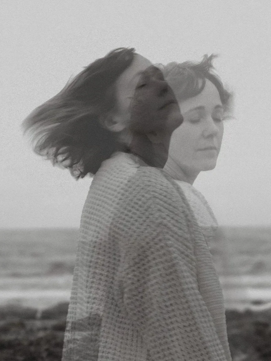 Black and white photo of two women with similar features, one with hair blowing in the wind, standing near the ocean, with a double exposure effect creating a layered appearance.
