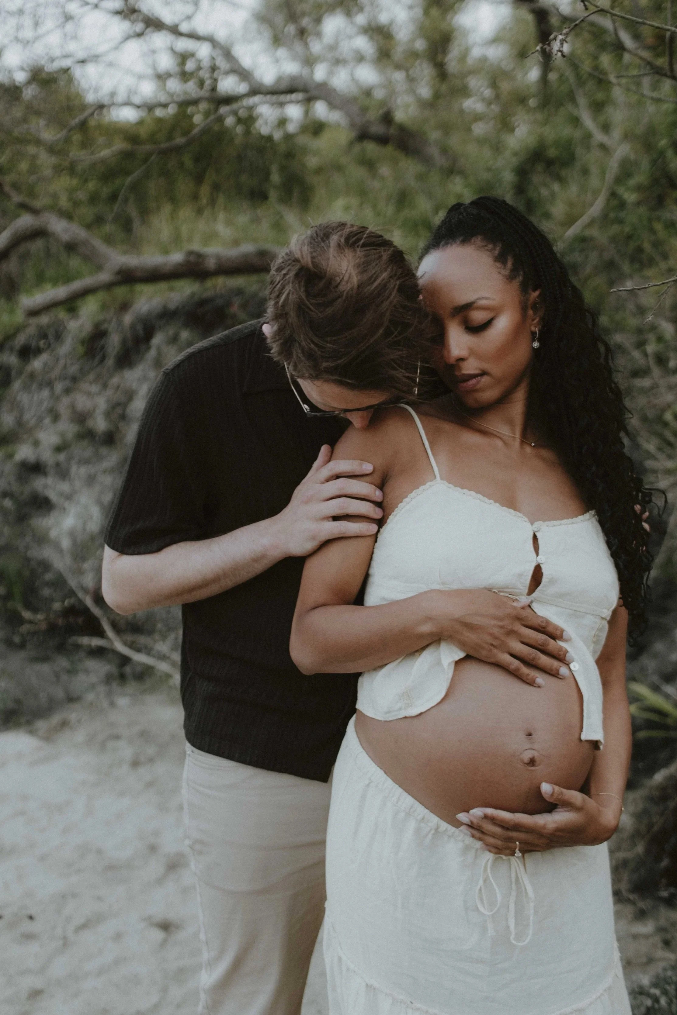 A pregnant woman with long curly black hair and a white top stands outdoors with a man with glasses and light skin, wearing a black shirt, placing his hand on her shoulder and leaning in close to her neck.