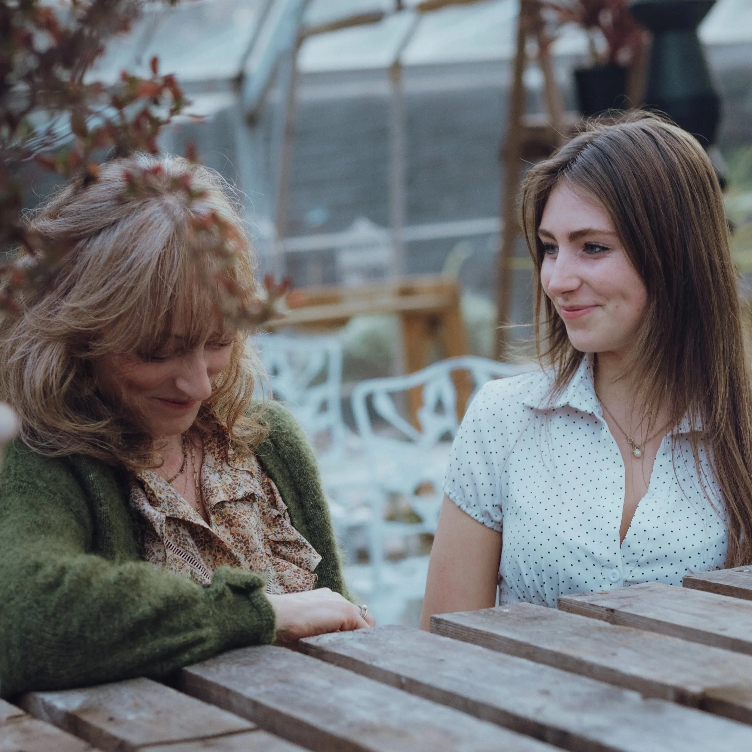 Two women sitting at a wooden table outdoors, smiling and engaging in a conversation. One has long brown hair and is wearing a white polka dot shirt, while the other has curly blonde hair and is wearing a green cardigan.