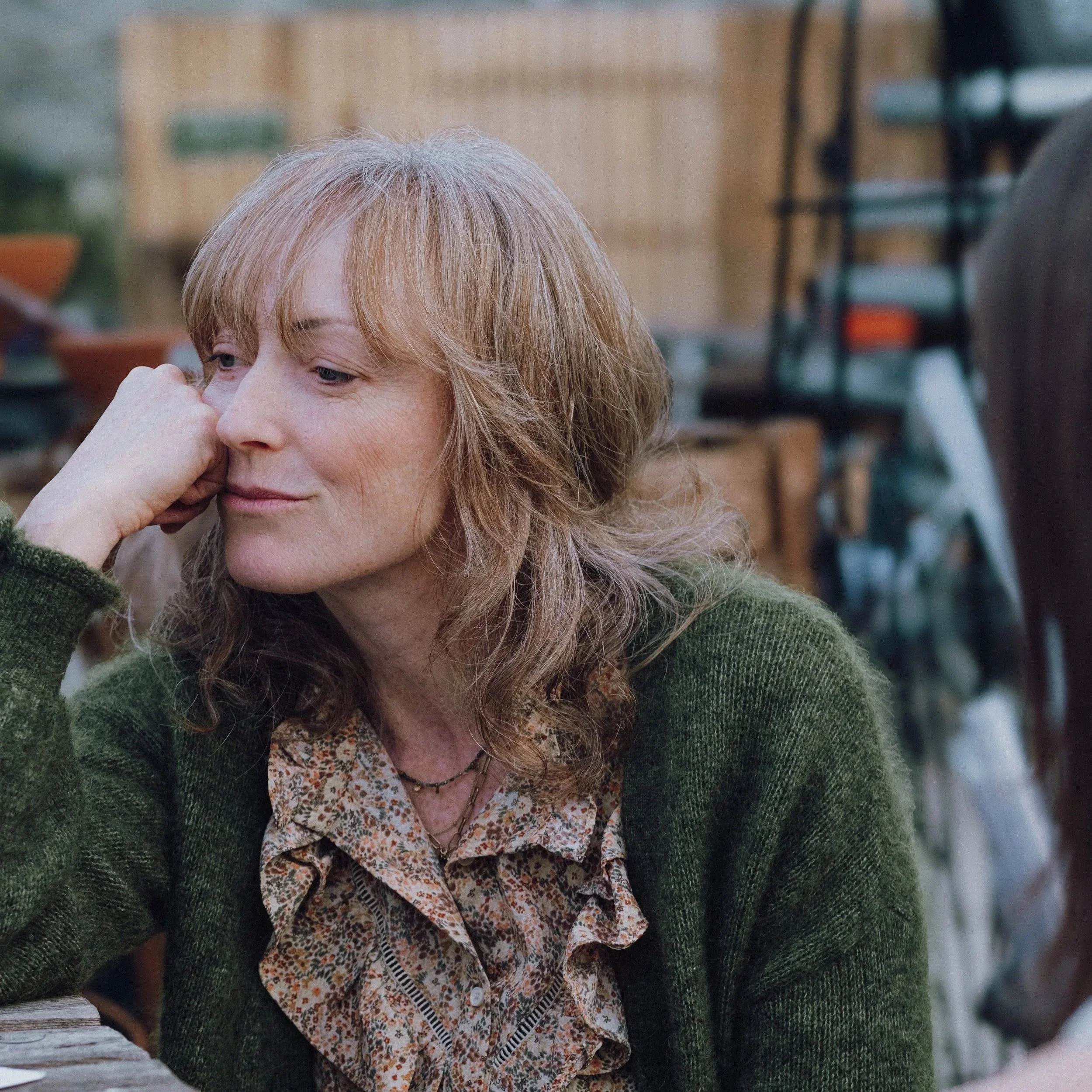A woman with shoulder-length blond hair resting her head on her hand, looking thoughtful, wearing a green sweater and a floral blouse, in an outdoor setting with gardening supplies in the background.