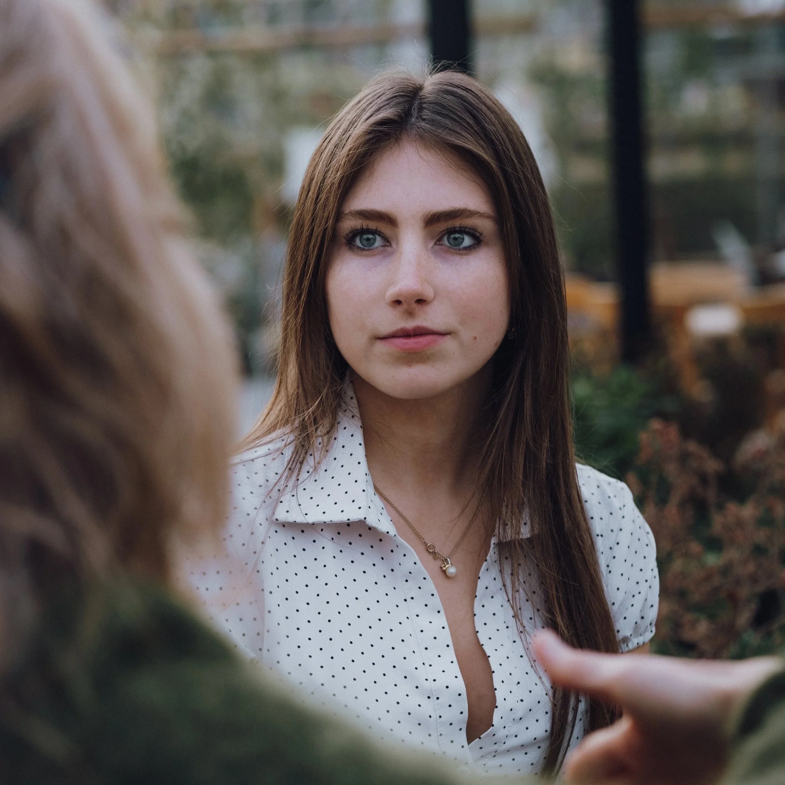 Close-up of a young woman with long brown hair and blue eyes, wearing a white shirt with black polka dots and a pearl necklace, listening attentively to someone out of frame.
