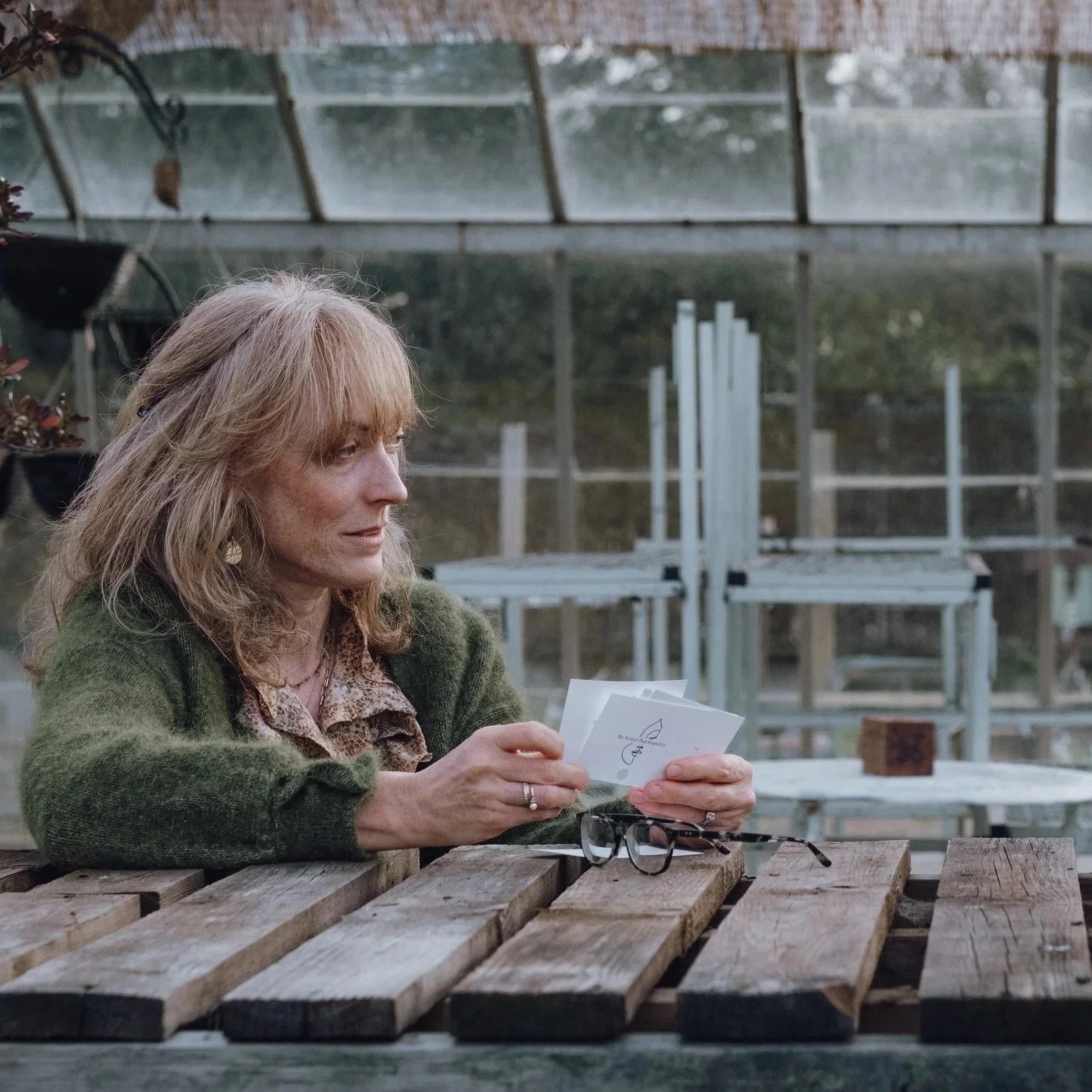 A woman with blonde hair wearing a green sweater and patterned blouse, sitting at a wooden table in a greenhouse, looking at a piece of paper.