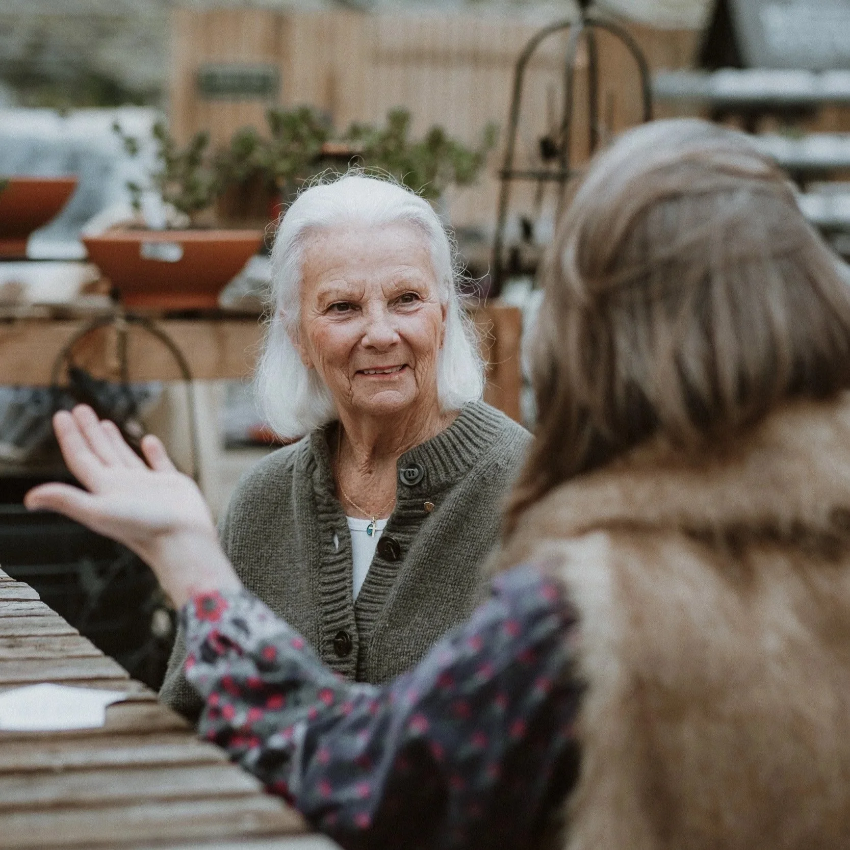 Two women are sitting at a wooden table in a garden or outdoor space, engaged in a conversation. One older woman with white hair is facing the camera, smiling, while the other woman with brown hair, wearing a fur vest and a patterned shirt, is seen f