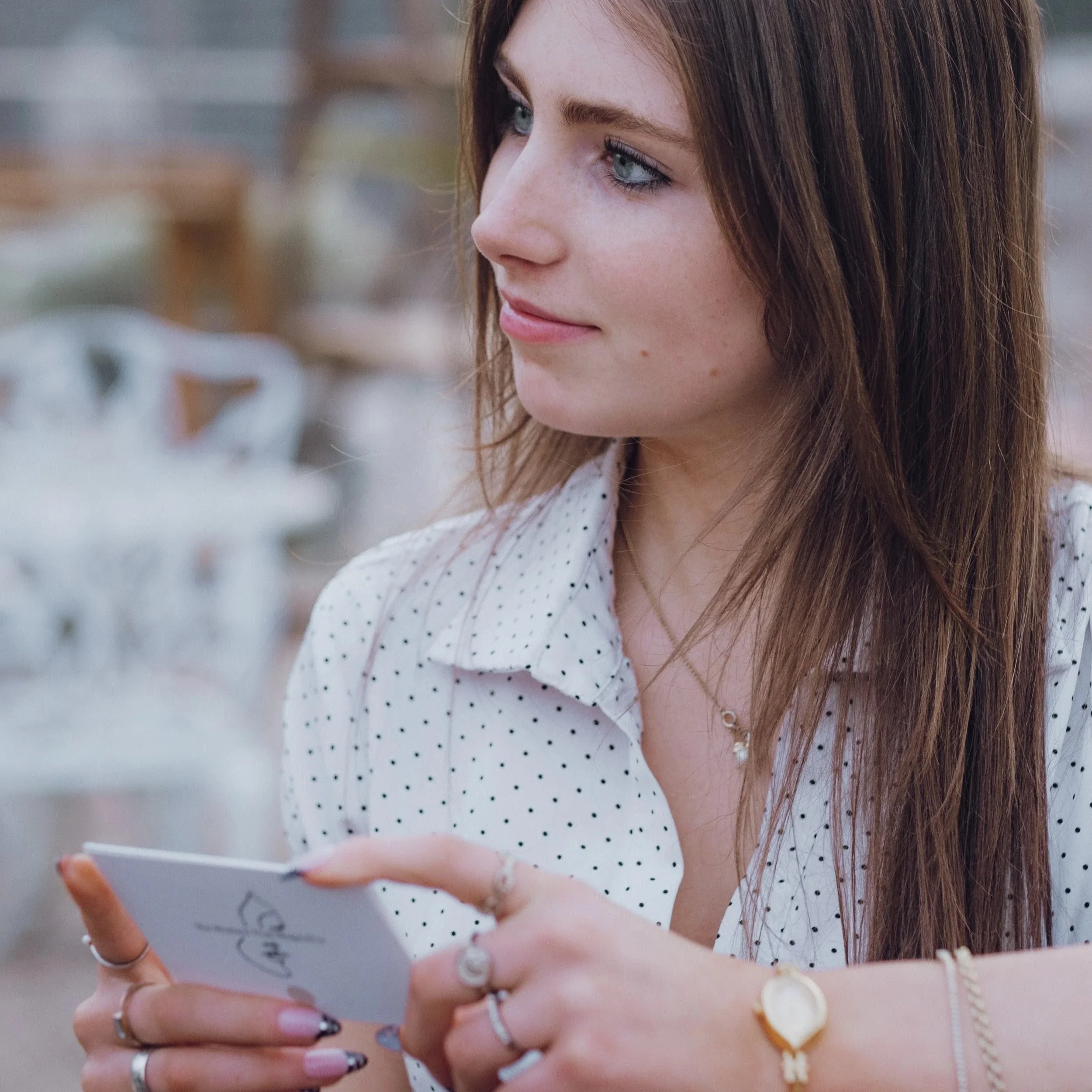 A young woman with long brown hair and blue eyes, wearing a white polka dot shirt, is holding a small notebook or card in her hand, looking to her left with a slight smile, at an outdoor cafe or similar setting.