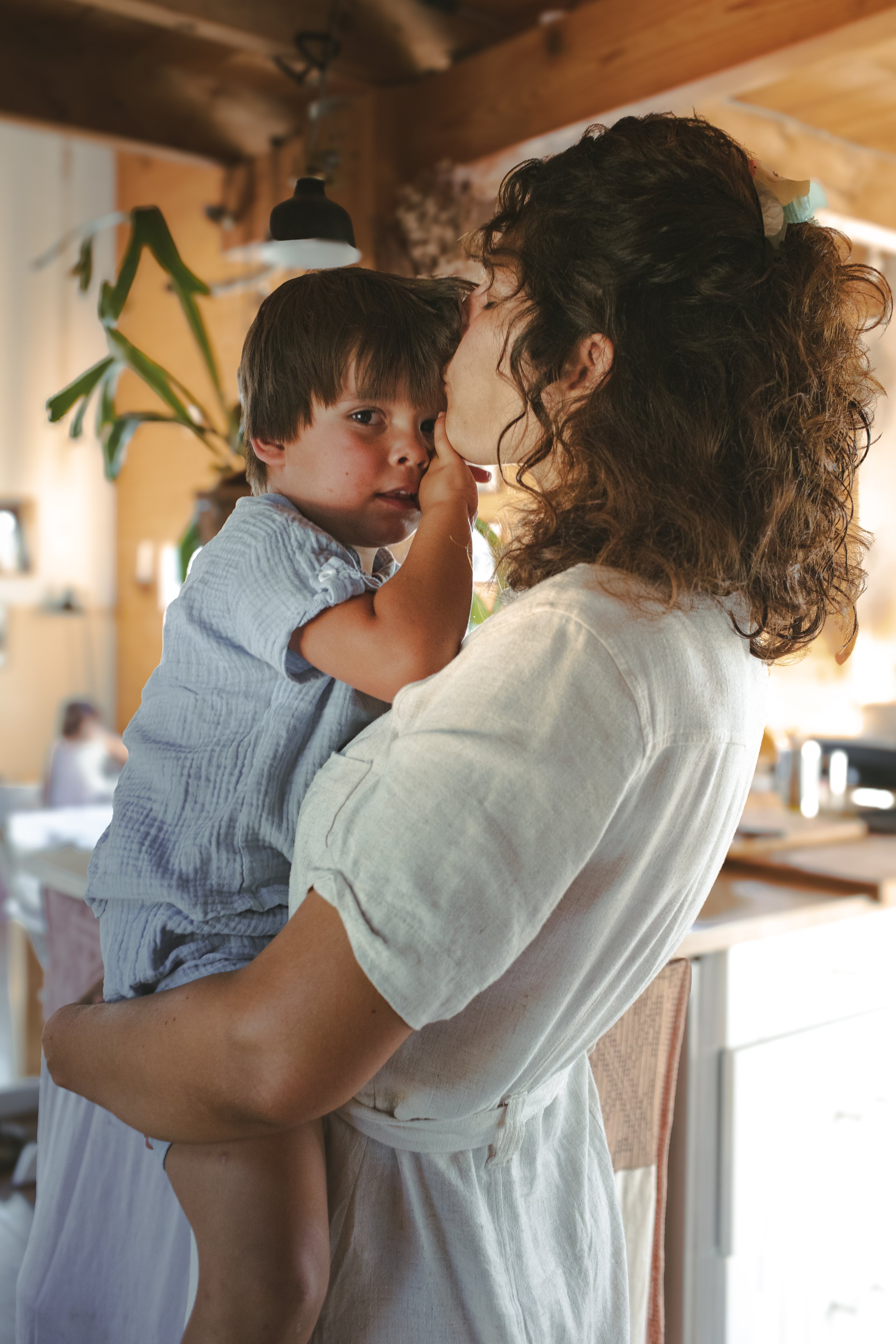 A woman with curly hair holding a young boy with straight brown hair, who is touching her face. They are indoors, with warm lighting and wooden accents.