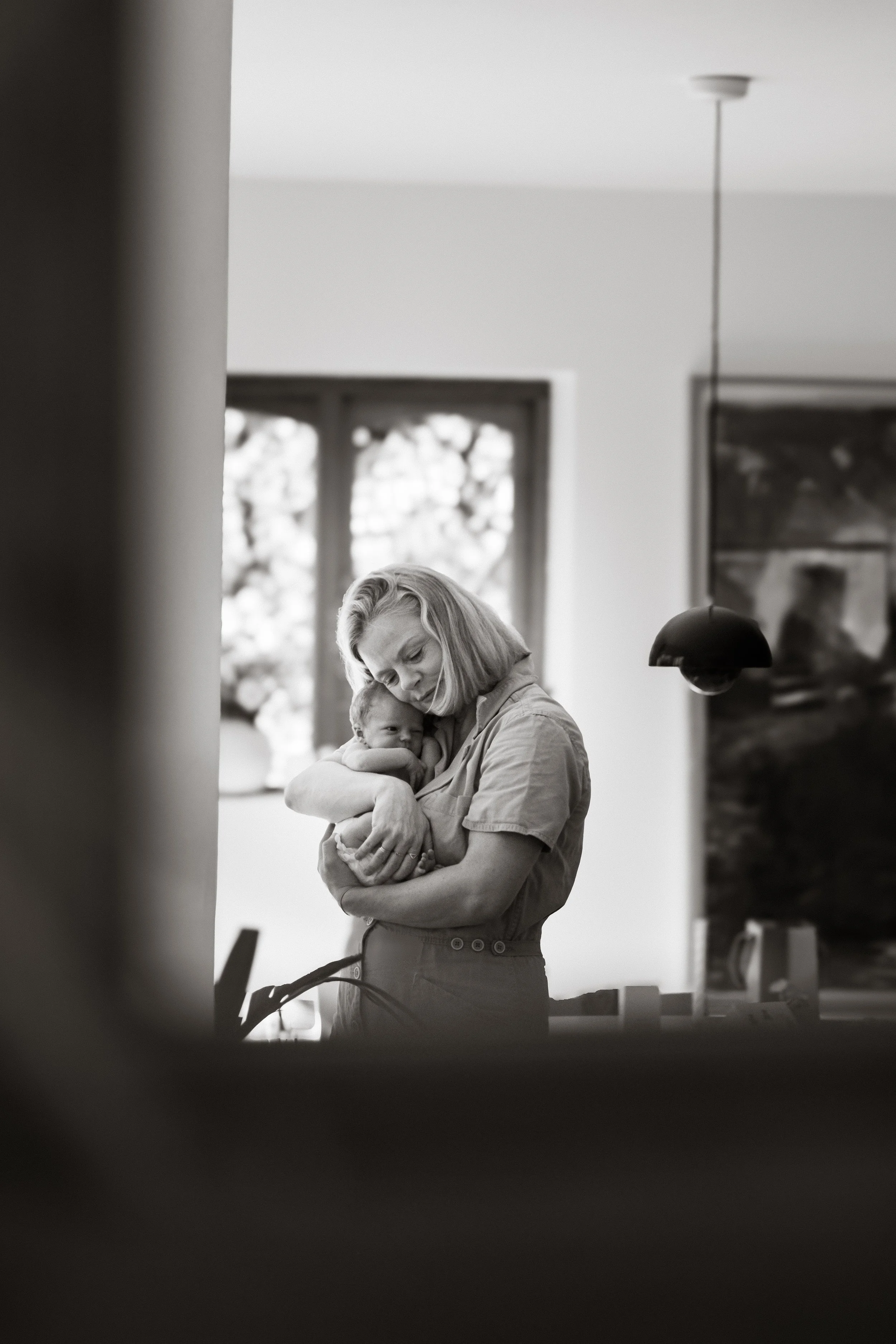 A woman holding a baby in a cozy room with a window in the background, captured in black and white.