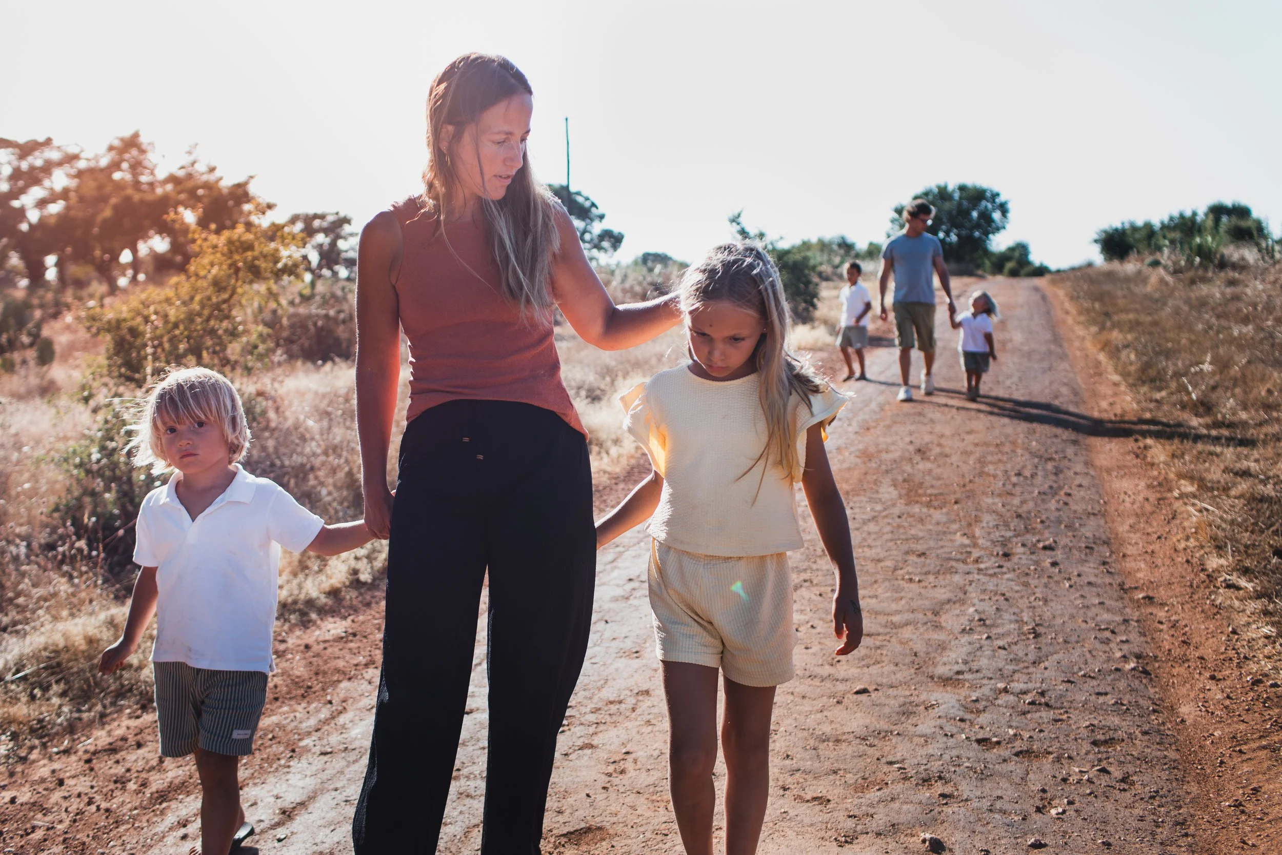 A woman walking outdoors on a dirt trail with four children, one holding her hand, in a dry, grassy landscape under a clear sky.