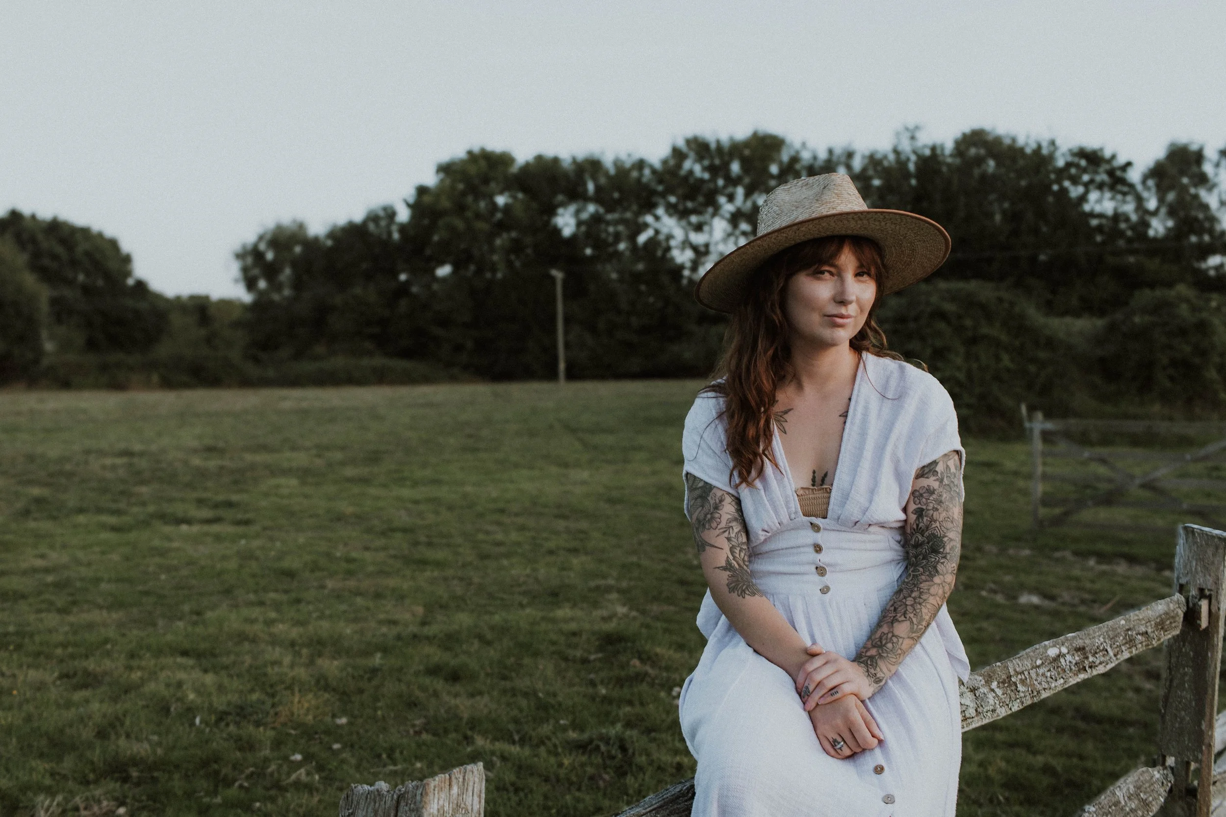 A young woman with long, wavy brown hair and tattoos on her arms, sitting outdoors on a rustic wooden fence. She is wearing a wide-brimmed straw hat, a light-colored button-up dress, and a beige top underneath. The background features a grassy field 