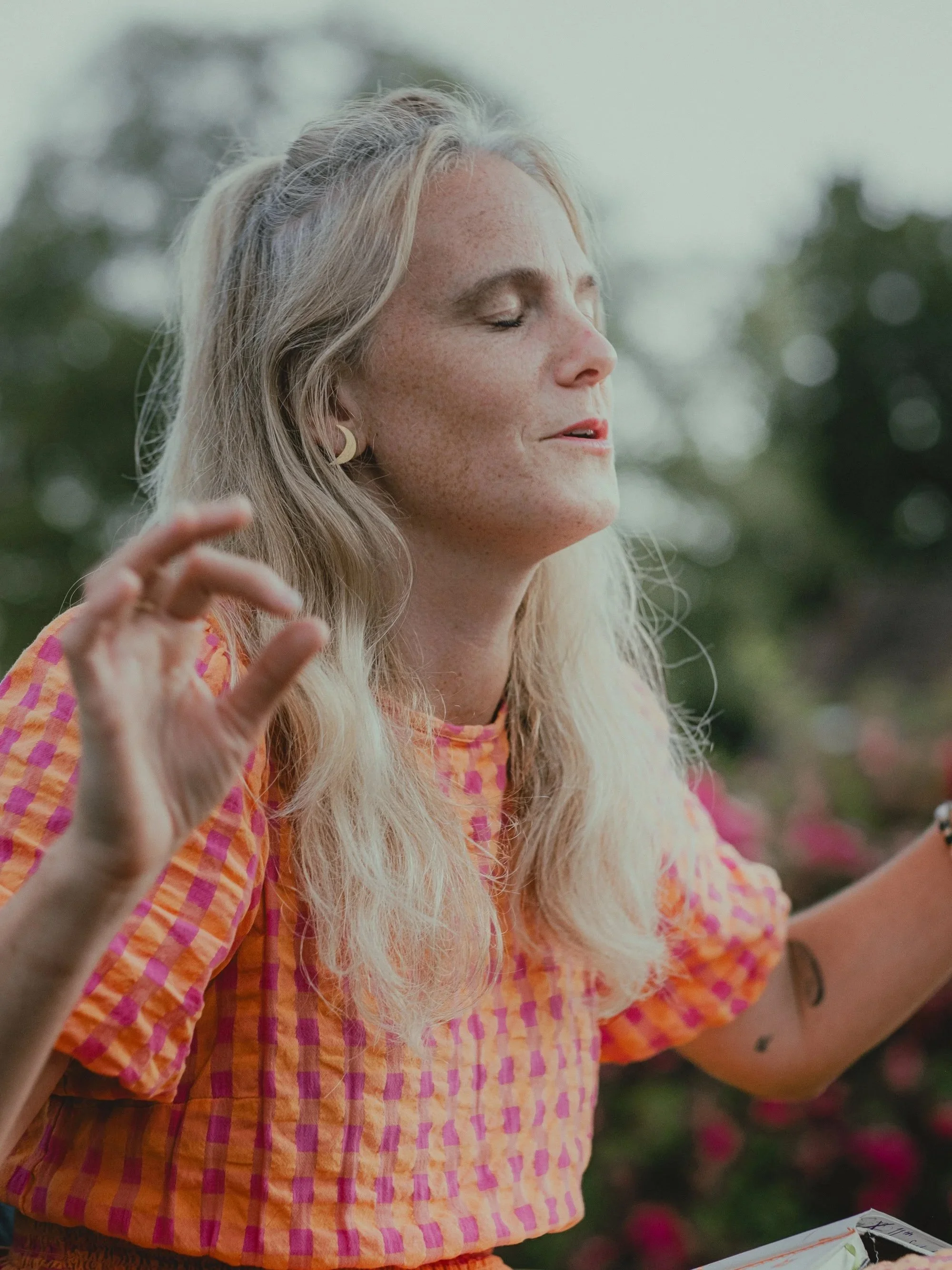 A woman with long, gray hair and a sun-kissed complexion, wearing a colorful, checkered dress, is outdoors with eyes closed, possibly in meditation or enjoying nature.