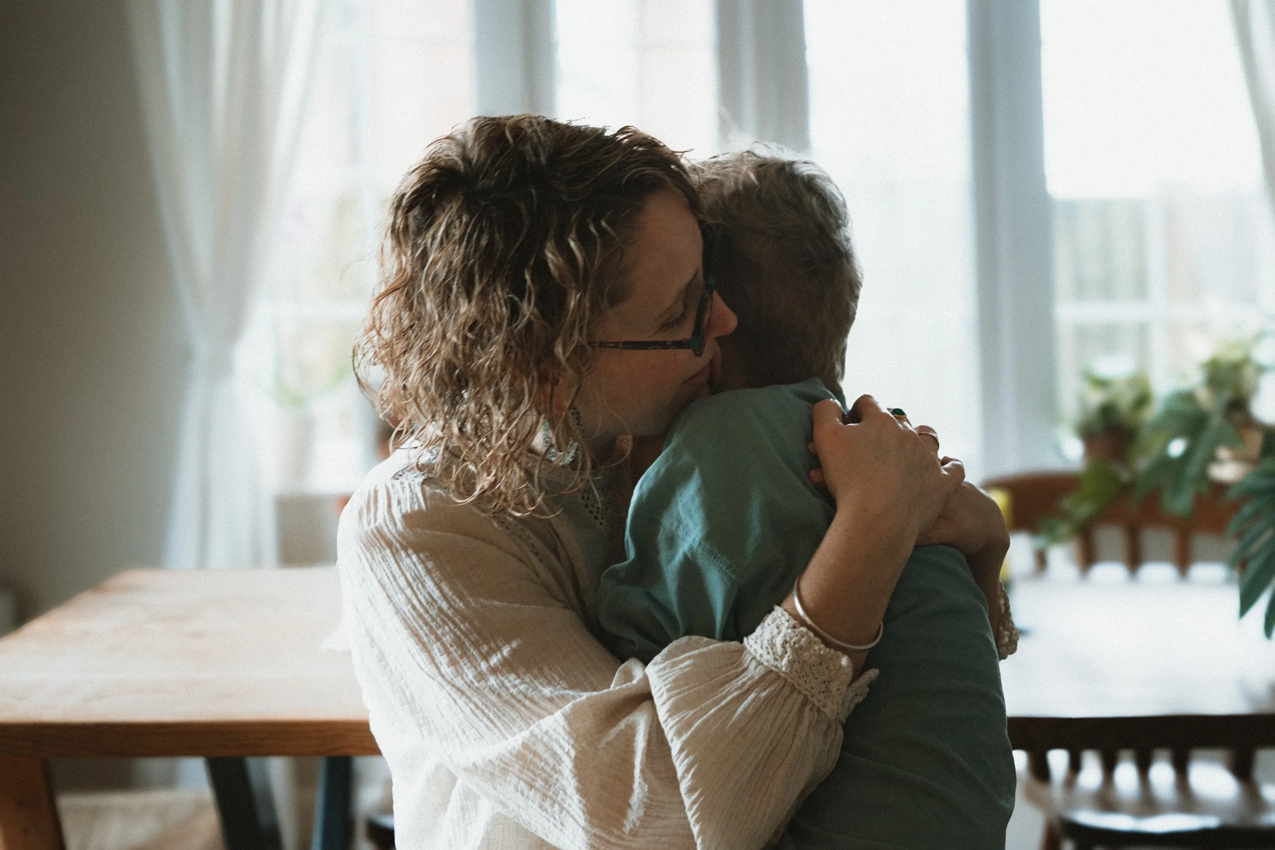 A woman with curly hair and glasses hugging a young boy with short hair in a room with natural light through large windows.