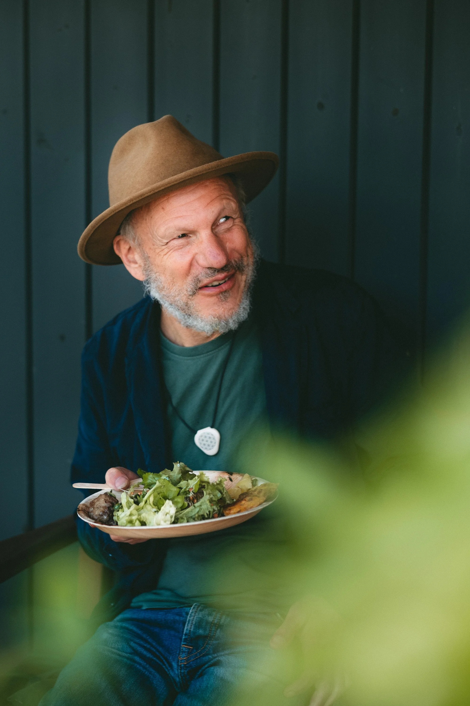 A smiling man with a gray beard wearing a brown hat and a dark blue jacket, holding a plate of salad and meat, sitting against a dark vertical wood-paneled background.