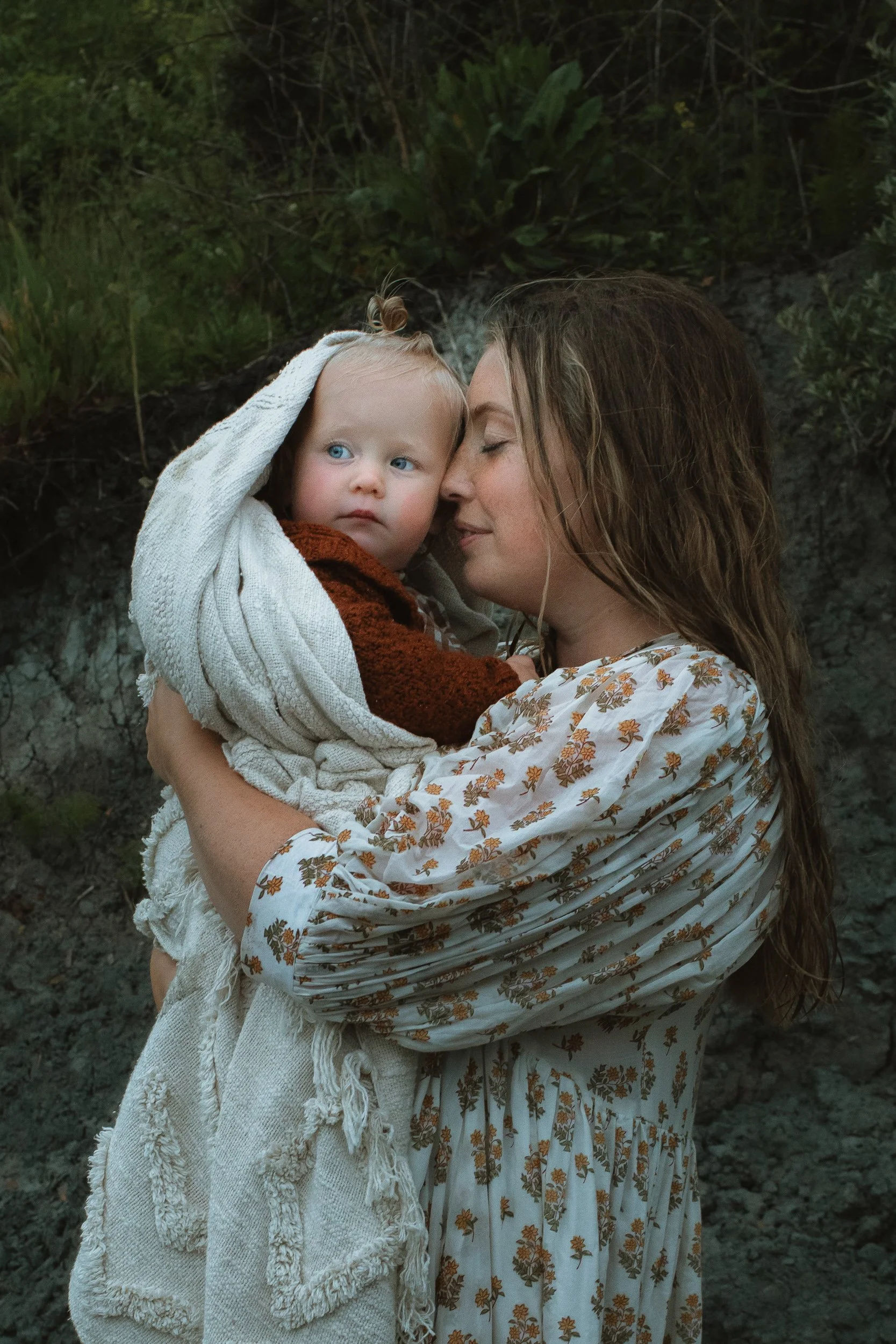 A woman holding a young child wrapped in a blanket outdoors, with greenery in the background.