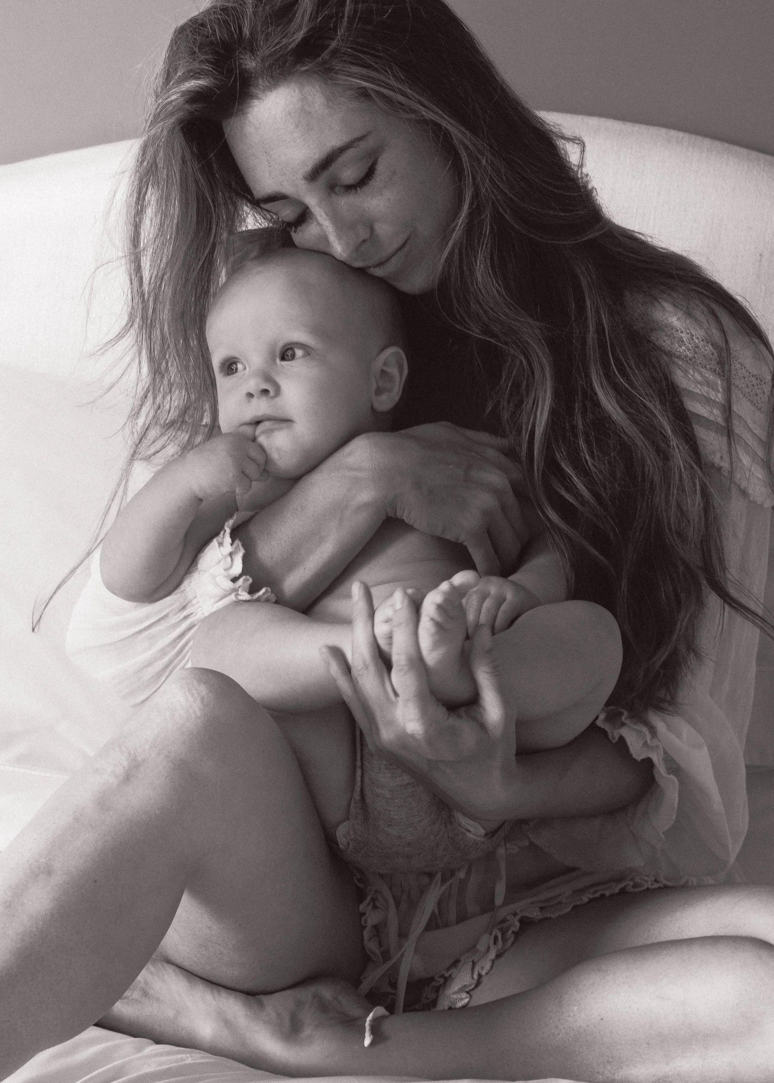 A woman cuddles a baby girl on a bed, both with long hair, in a black-and-white photograph.