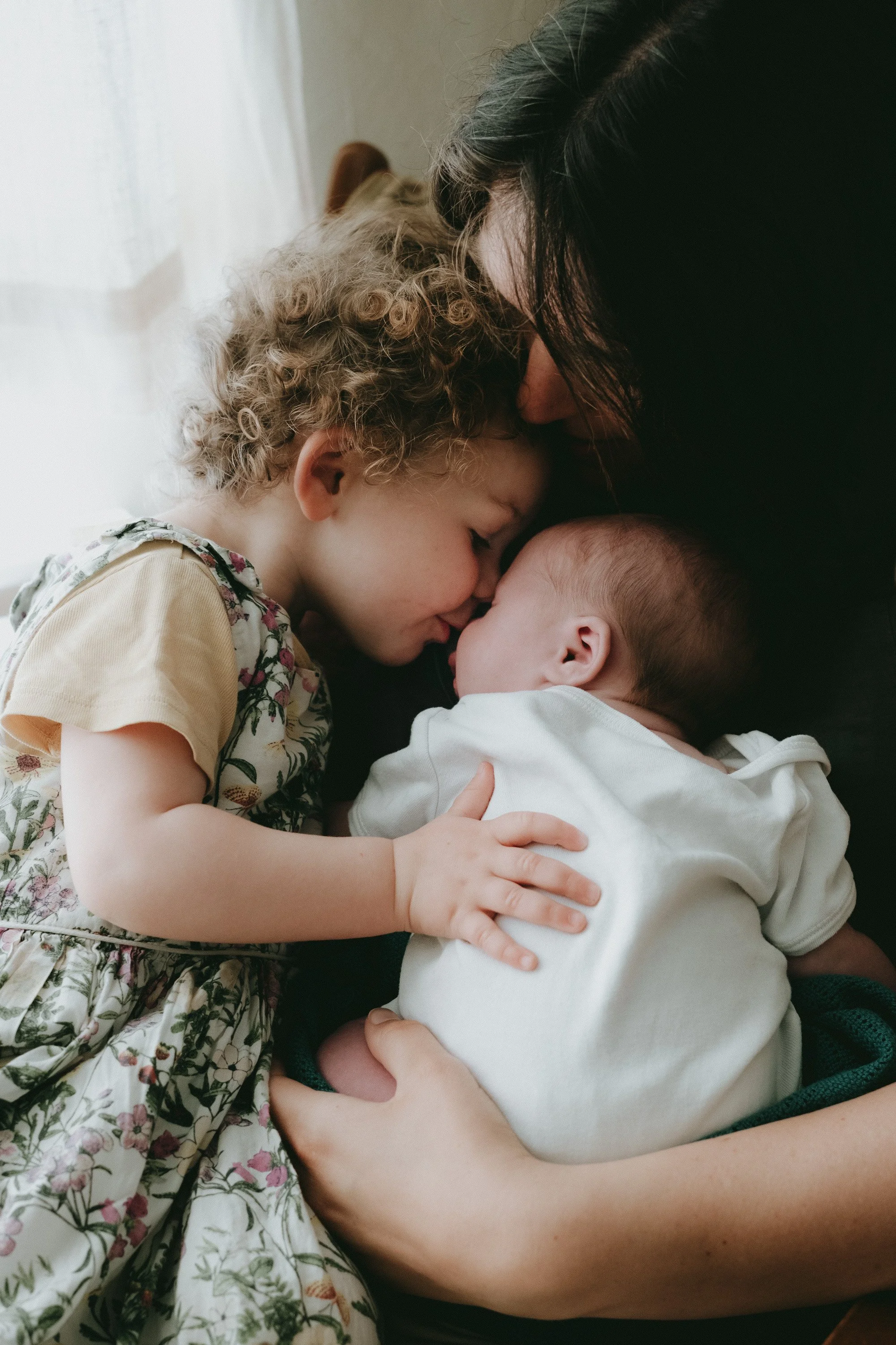 A woman and two children sharing a tender moment, with the woman gently pressing her forehead against a baby’s head and the older child holding the baby, all appearing close and affectionate.