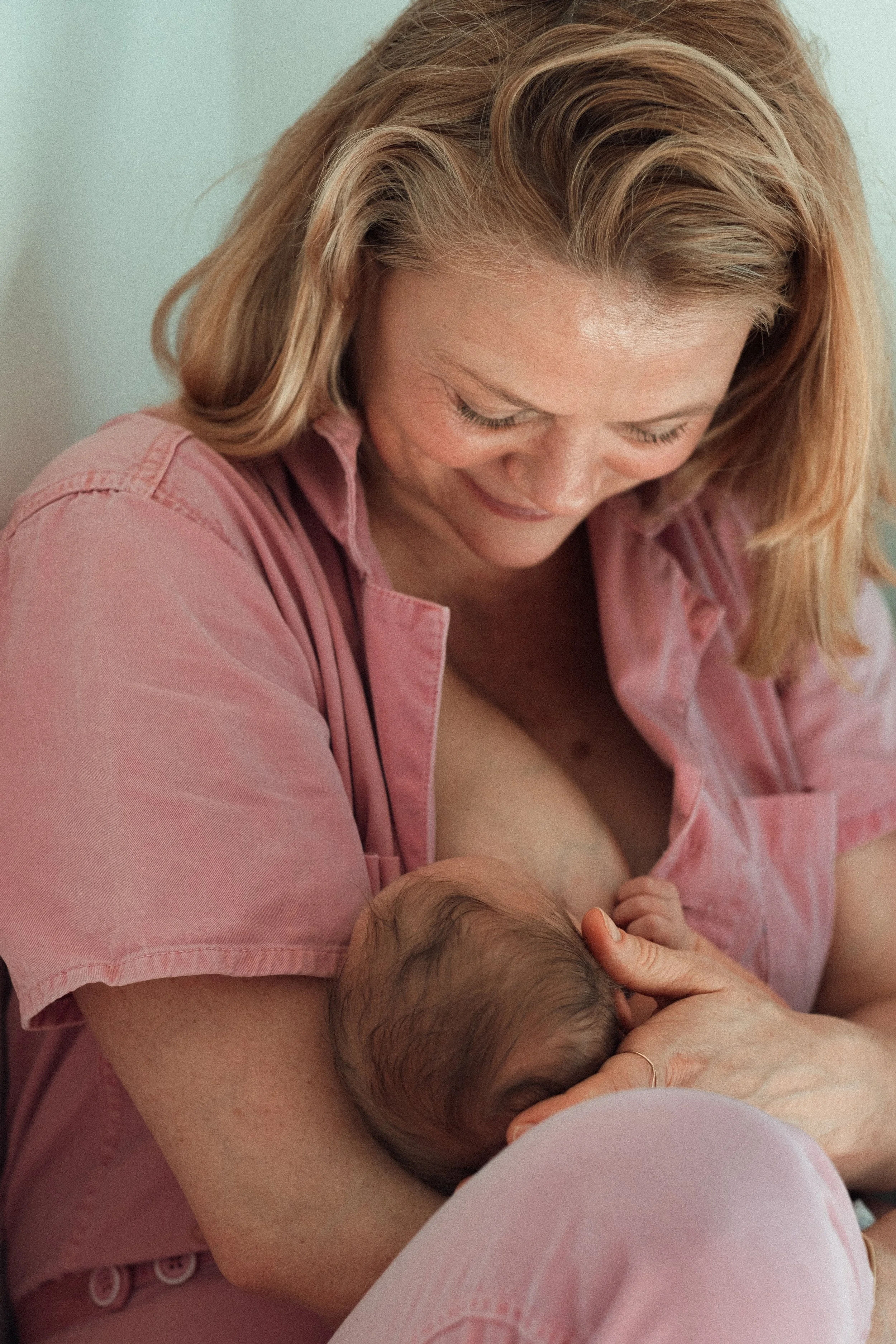 A woman with blonde hair wearing a pink shirt breastfeeding a newborn baby.