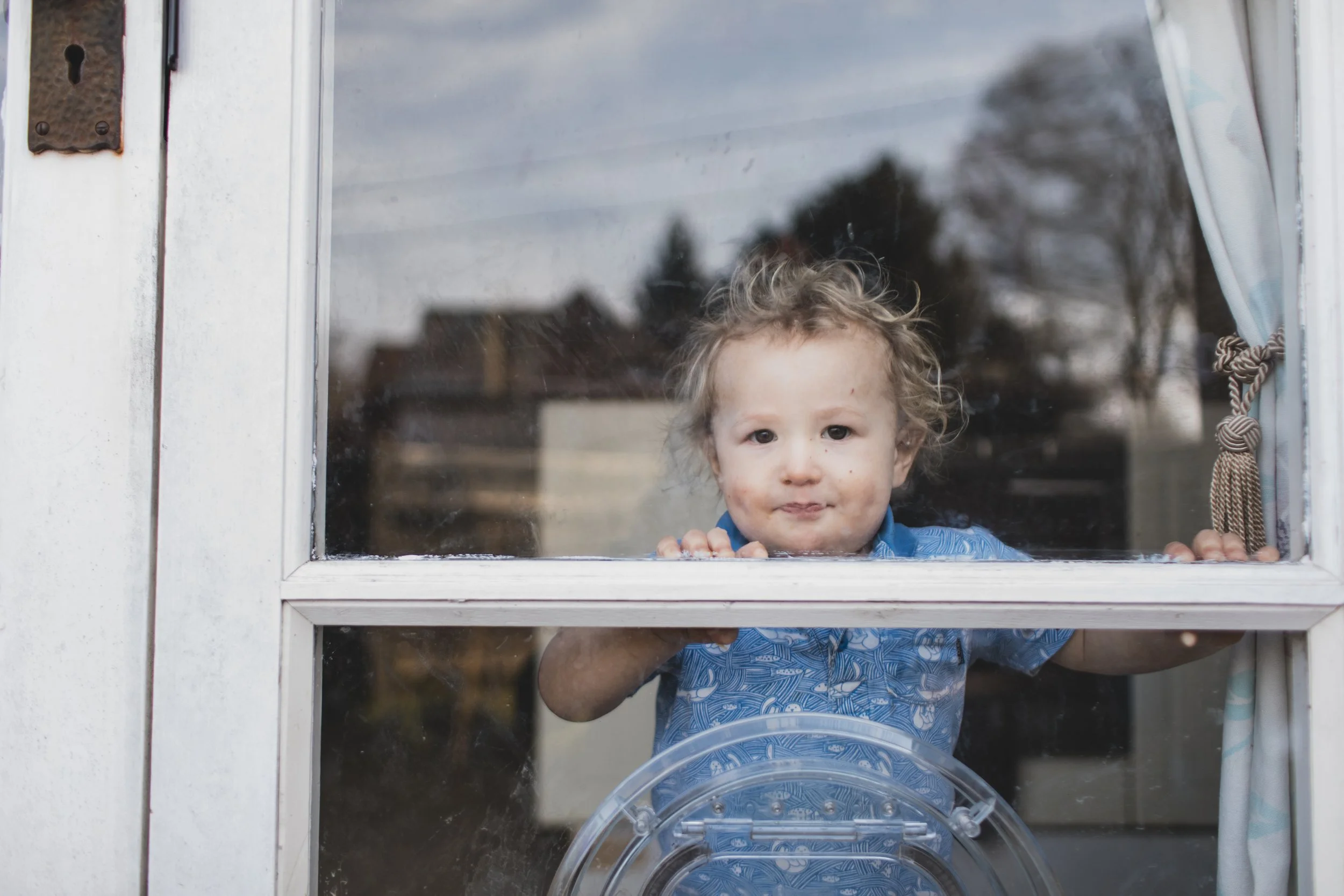 A young child with curly hair looking through a window with a curious expression.