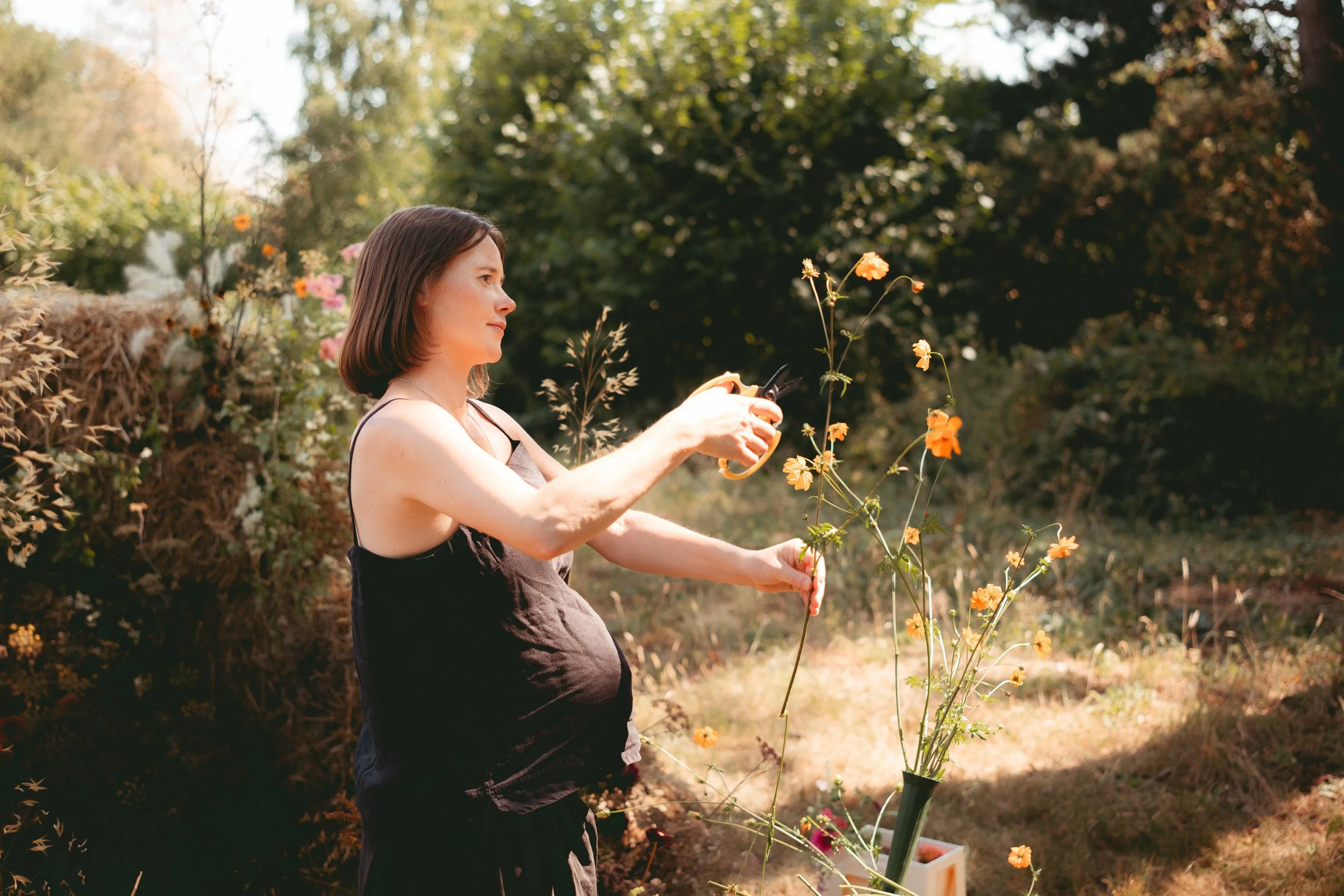 A woman with short brown hair wearing a black sleeveless top and apron, watering orange flowers in her garden during daytime with sunlight filtering through trees.
