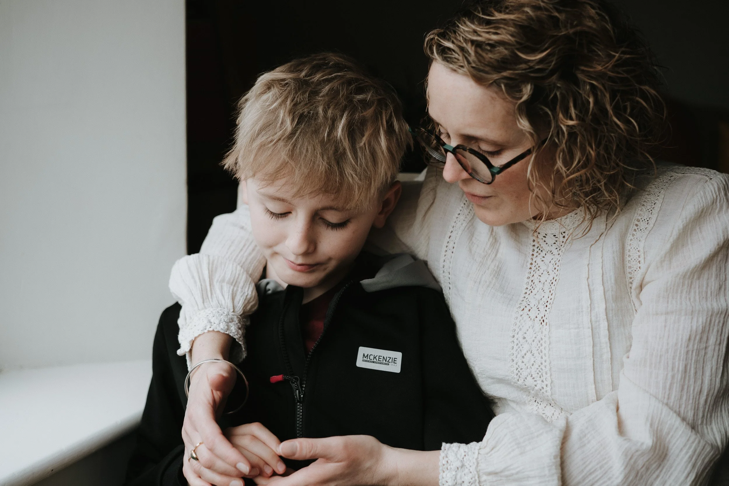 A woman with curly hair and glasses holding hands with a boy with closed eyes and blonde hair, both looking down at something in the woman's hands and sitting closely together near a window.