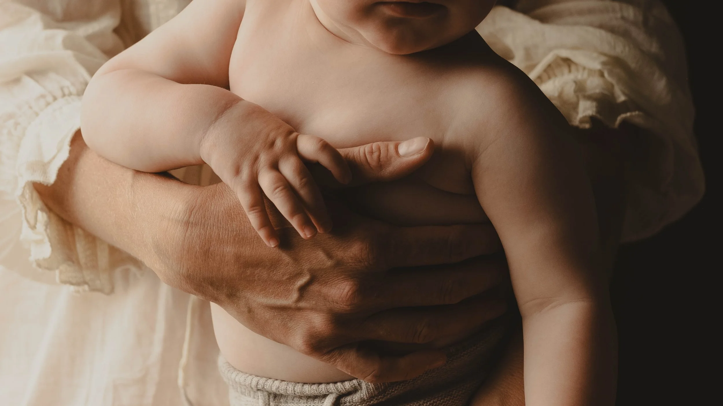 A small child's hand gently resting on an adult's hand, both of them are partially clothed and seated, with soft lighting highlighting their skin tones.