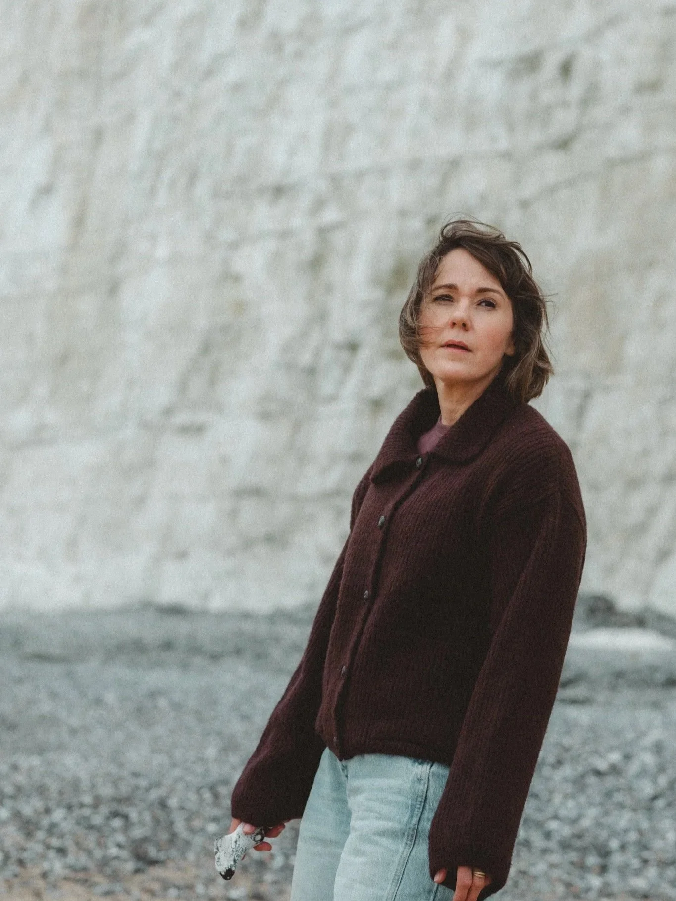 A woman with short brown hair wearing a dark brown coat and light blue jeans stands outdoors against a large gray stone wall. She is holding a pair of sunglasses in her right hand and gazes into the distance.