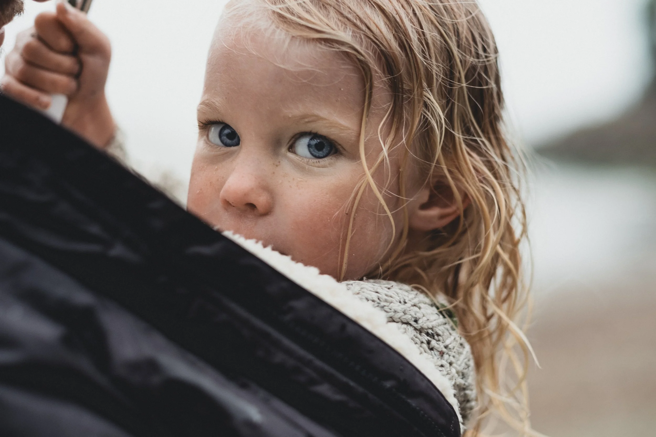 Close-up of a young girl with blond curly hair and blue eyes, looking over her shoulder at the camera, with a cloudy outdoor background.