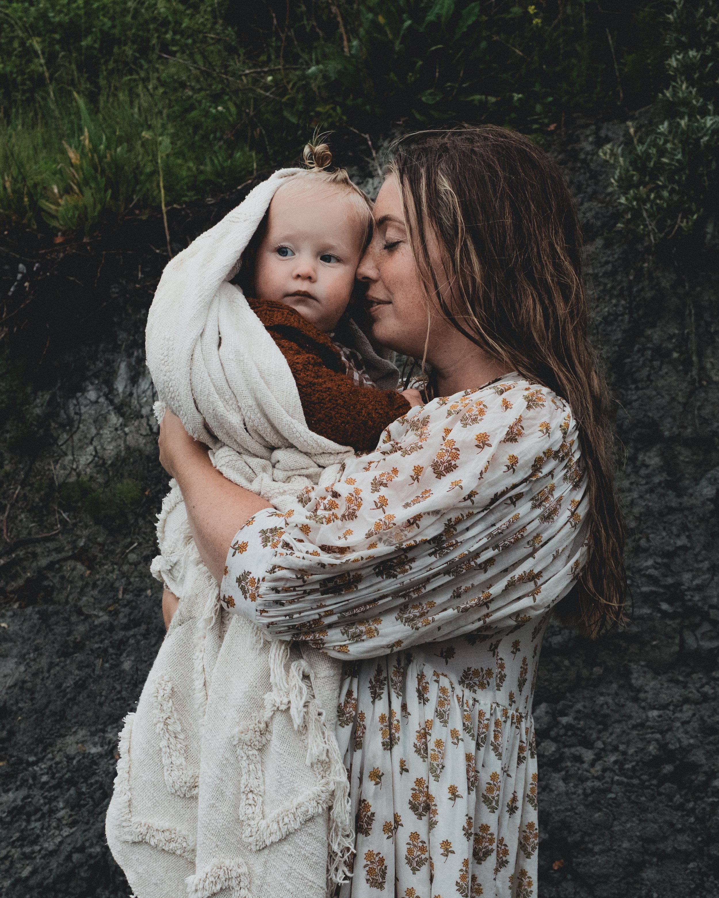 A woman with long brown hair holding a young child wrapped in a cream-colored blanket, outdoors with green foliage in the background.
