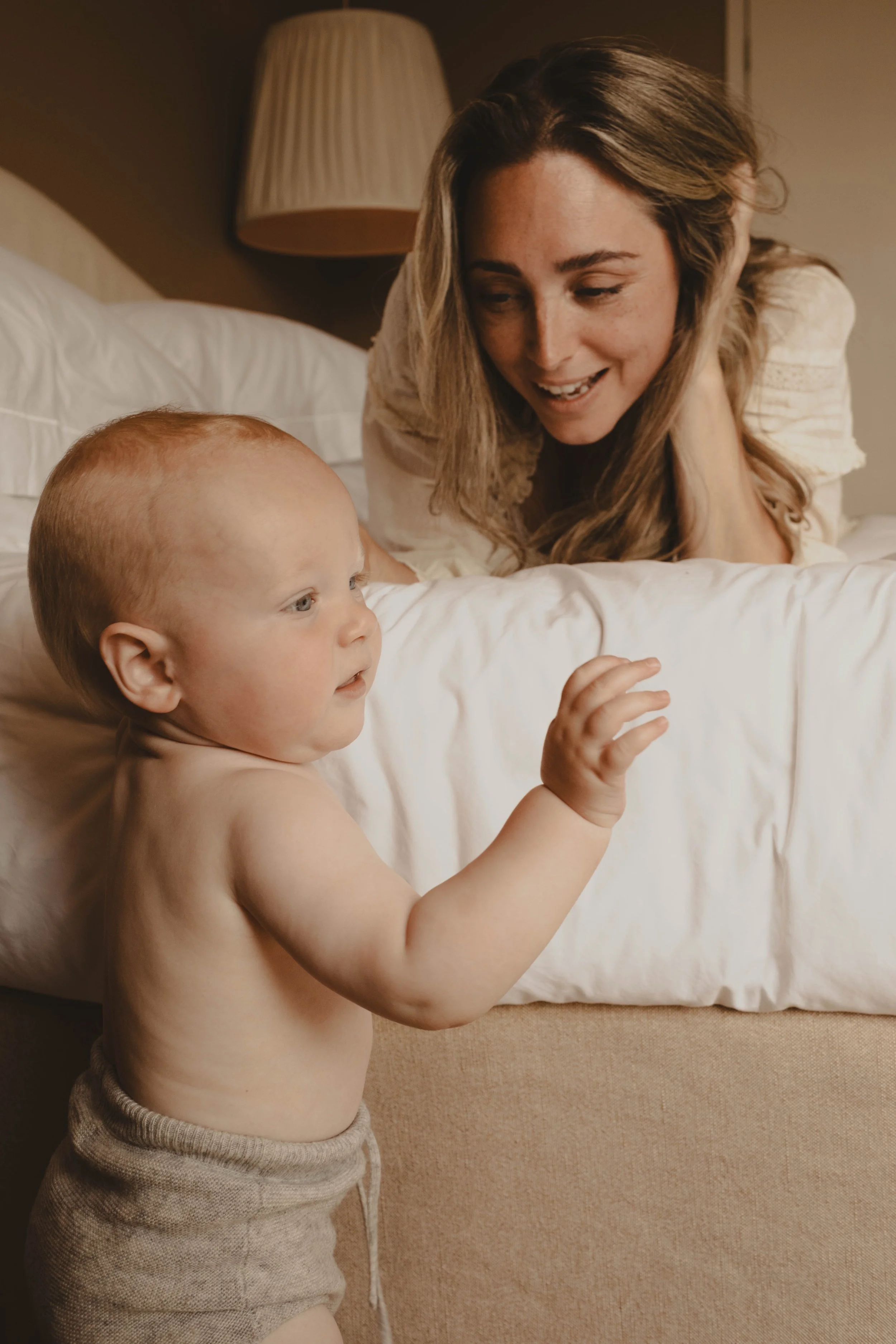 A woman and a toddler boy with a shaved head and no shirt are interacting in a bedroom. The woman is leaning over the bed, smiling at the boy, who is standing beside the bed.