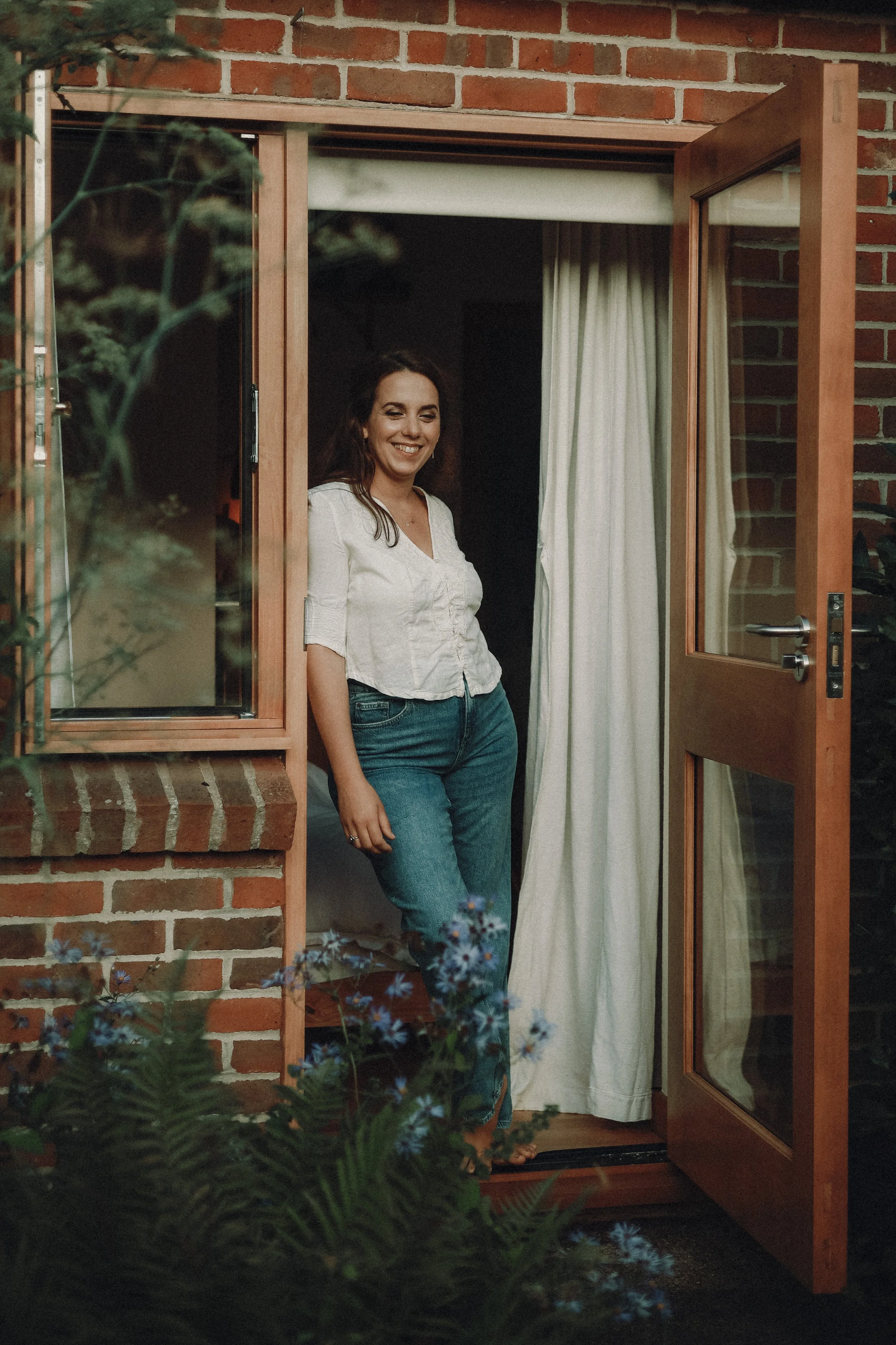 A woman with dark hair and a smile, wearing a white blouse and jeans, stands at an open wooden window of a brick house, with a curtain behind her. There are plants and flowers in the foreground.