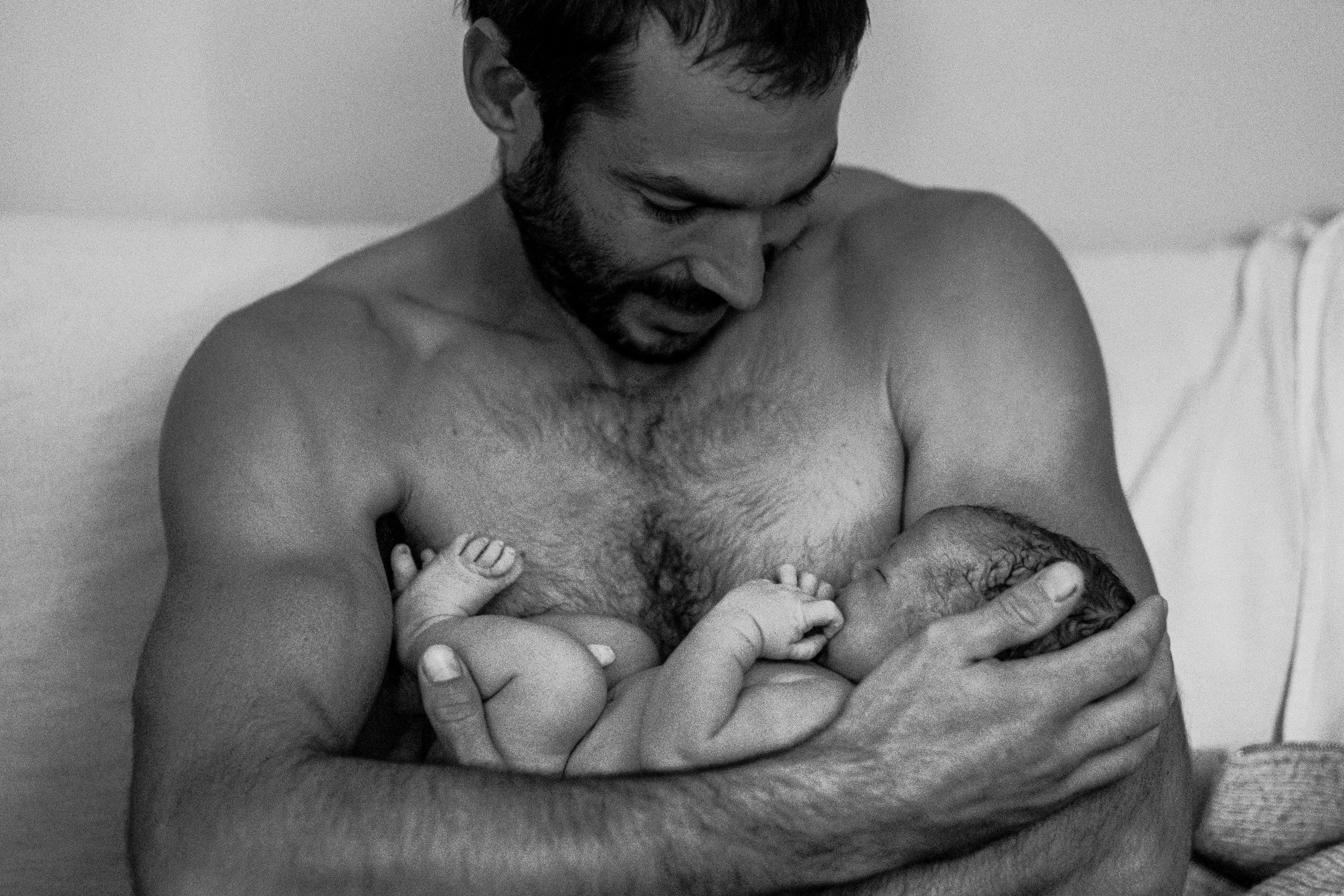 Black and white photo of a shirtless man with a beard breastfeeding a newborn baby at home, sitting on a couch.