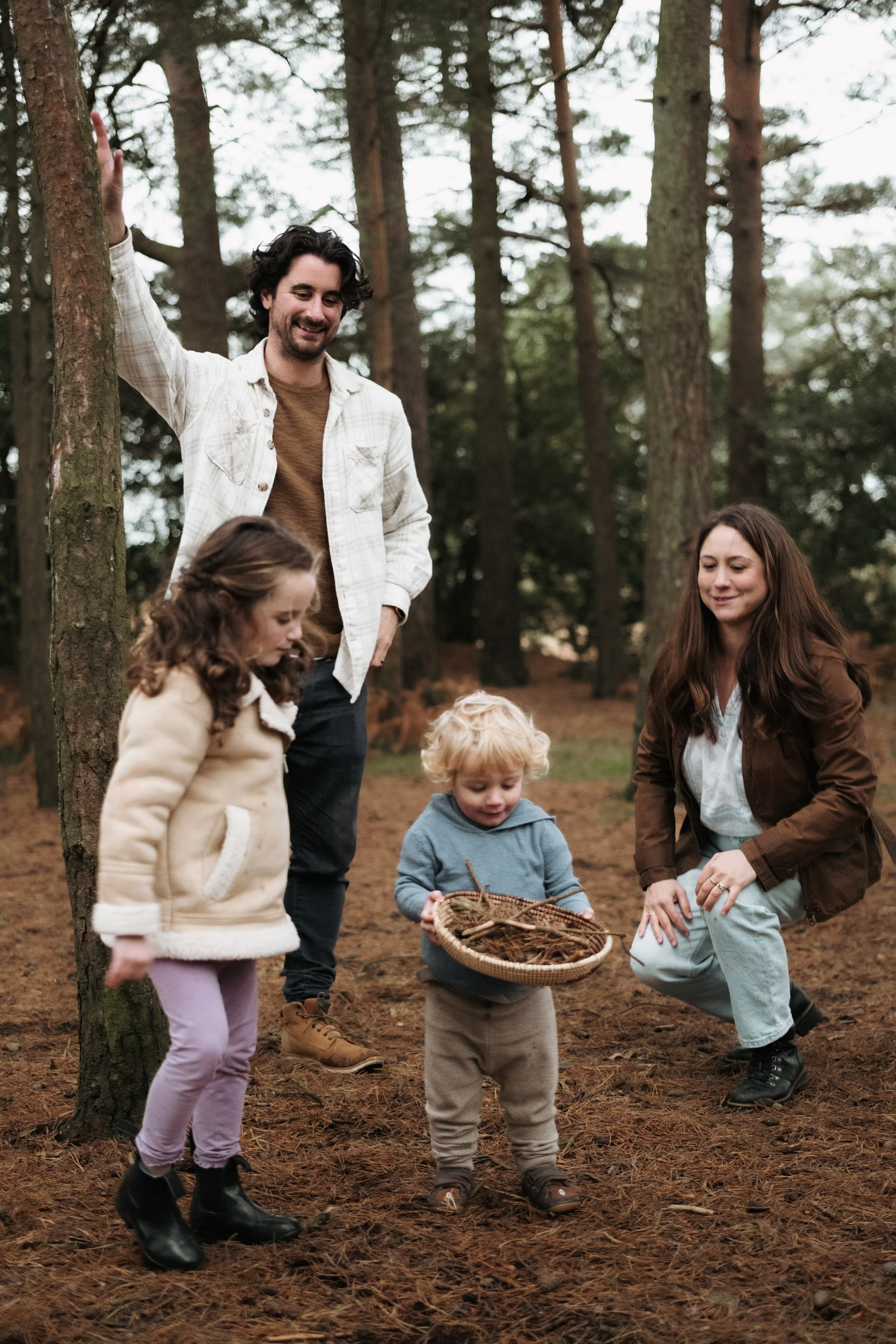 A family collecting pinecones in a forest, with two adults and three children, surrounded by tall trees and pine needles on the ground.