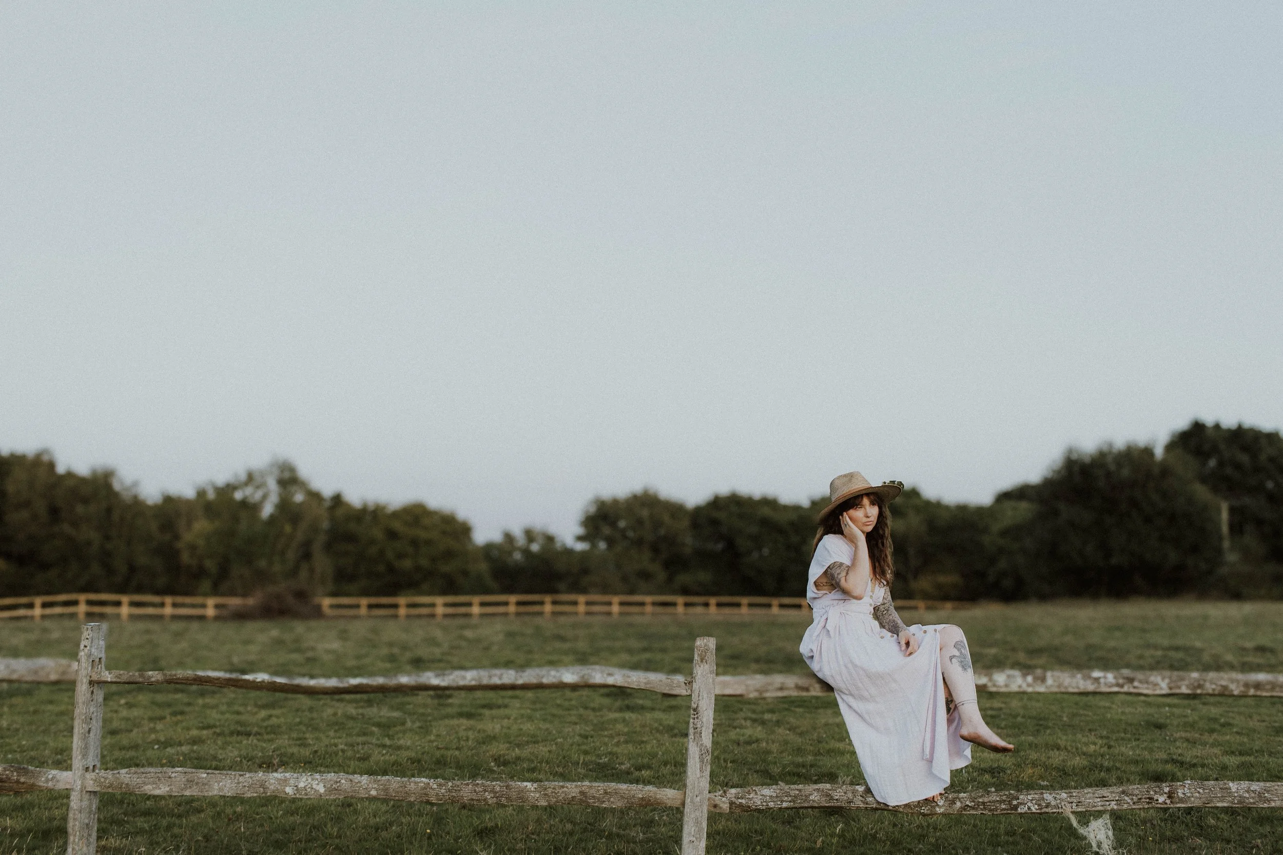 A woman with long hair wearing a white dress and large hat sitting on a wooden fence in a grassy field with trees in the background.