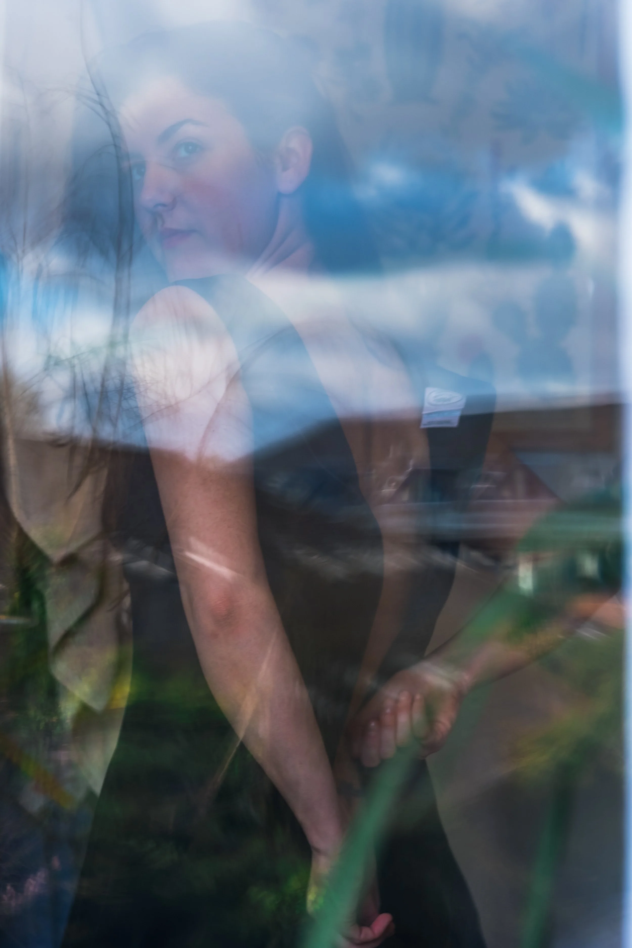 A woman in a black dress looks through a reflective surface, with trees and a blue sky visible in the reflection.