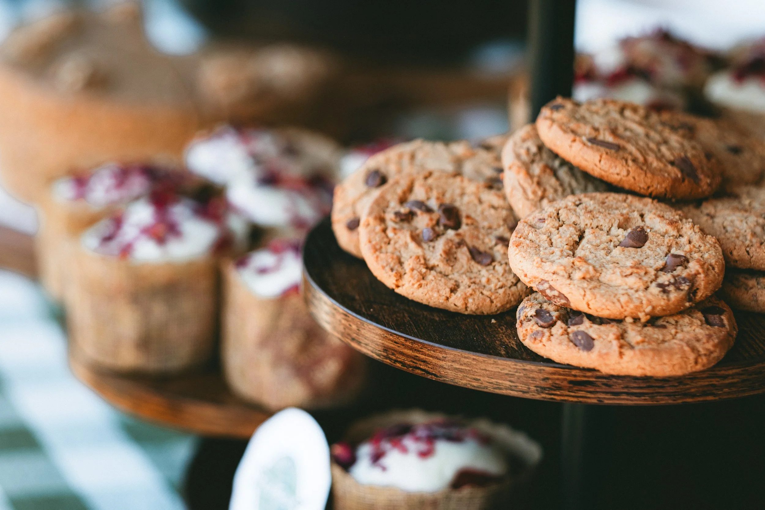 Close-up of chocolate chip cookies on a dark wooden plate, with blurred background of cupcakes.