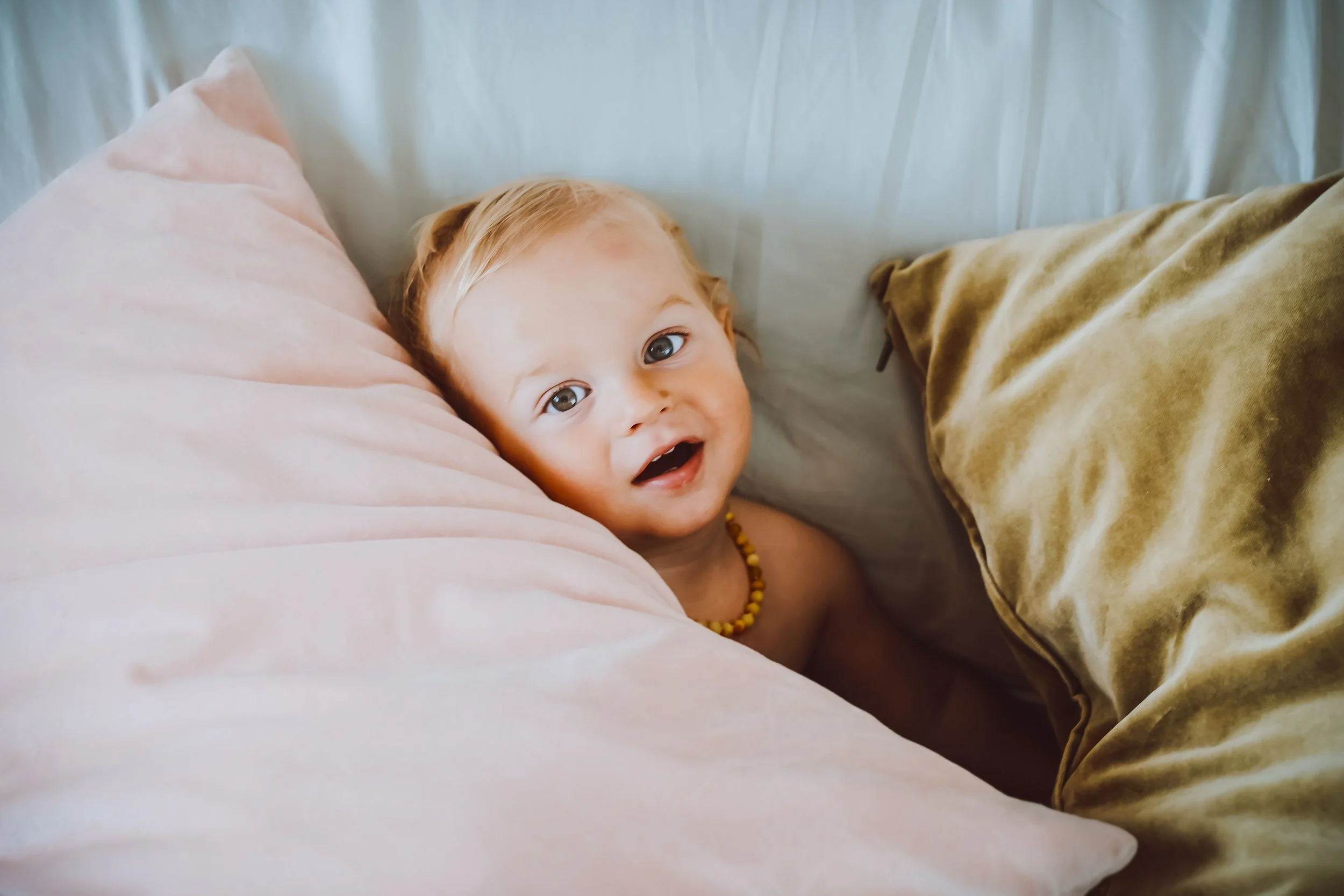 A young child with red hair, lying in bed between pink and tan cushions, looking up and smiling.