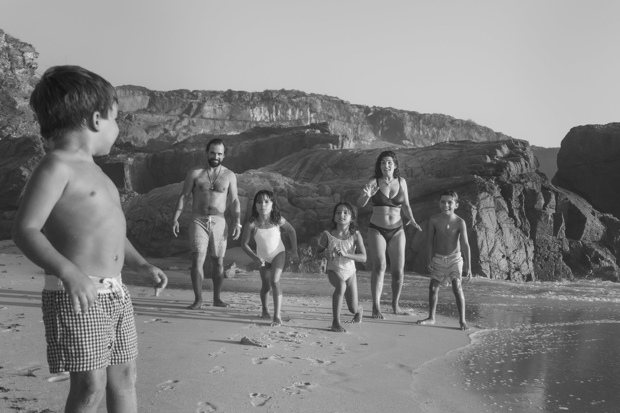 A group of five children and two adults on a beach, with rocky cliffs in the background, all dressed in swimwear, walking along the shoreline.