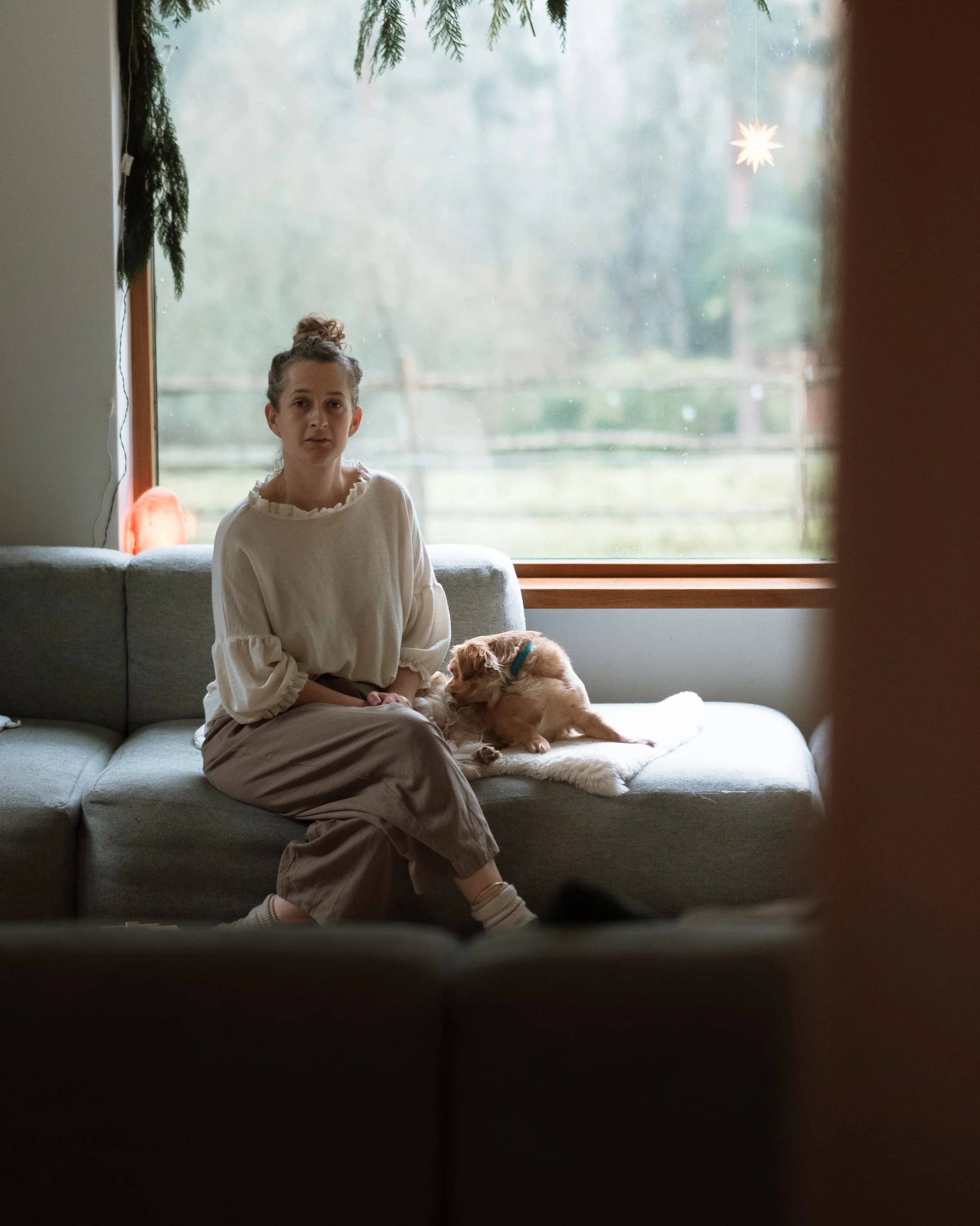 A woman sitting on a sofa with a small dog on a blanket next to her, in a room with large window showing an outdoor view.