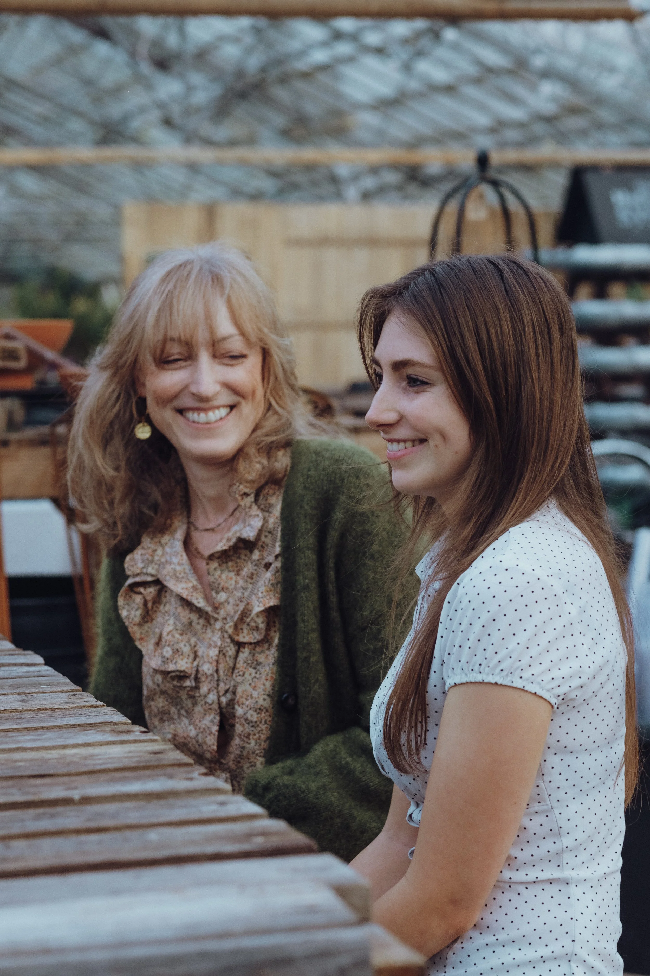 Two women sitting at a wooden table, smiling and talking in a greenhouse or garden setting.