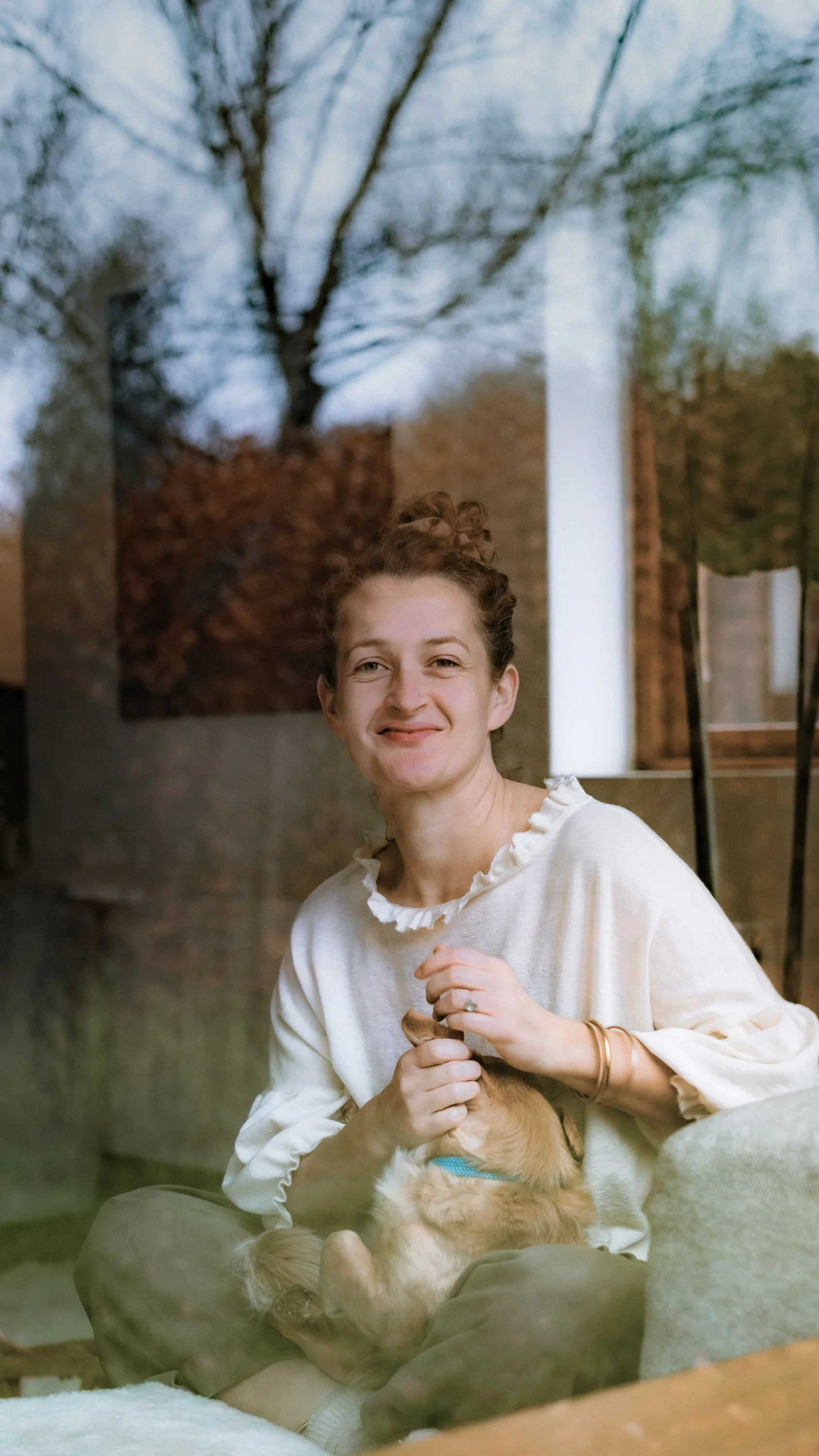 A woman with light skin and curly hair tied up in a bun, smiling and sitting on a sofa holding a small dog with light brown fur and a blue collar.