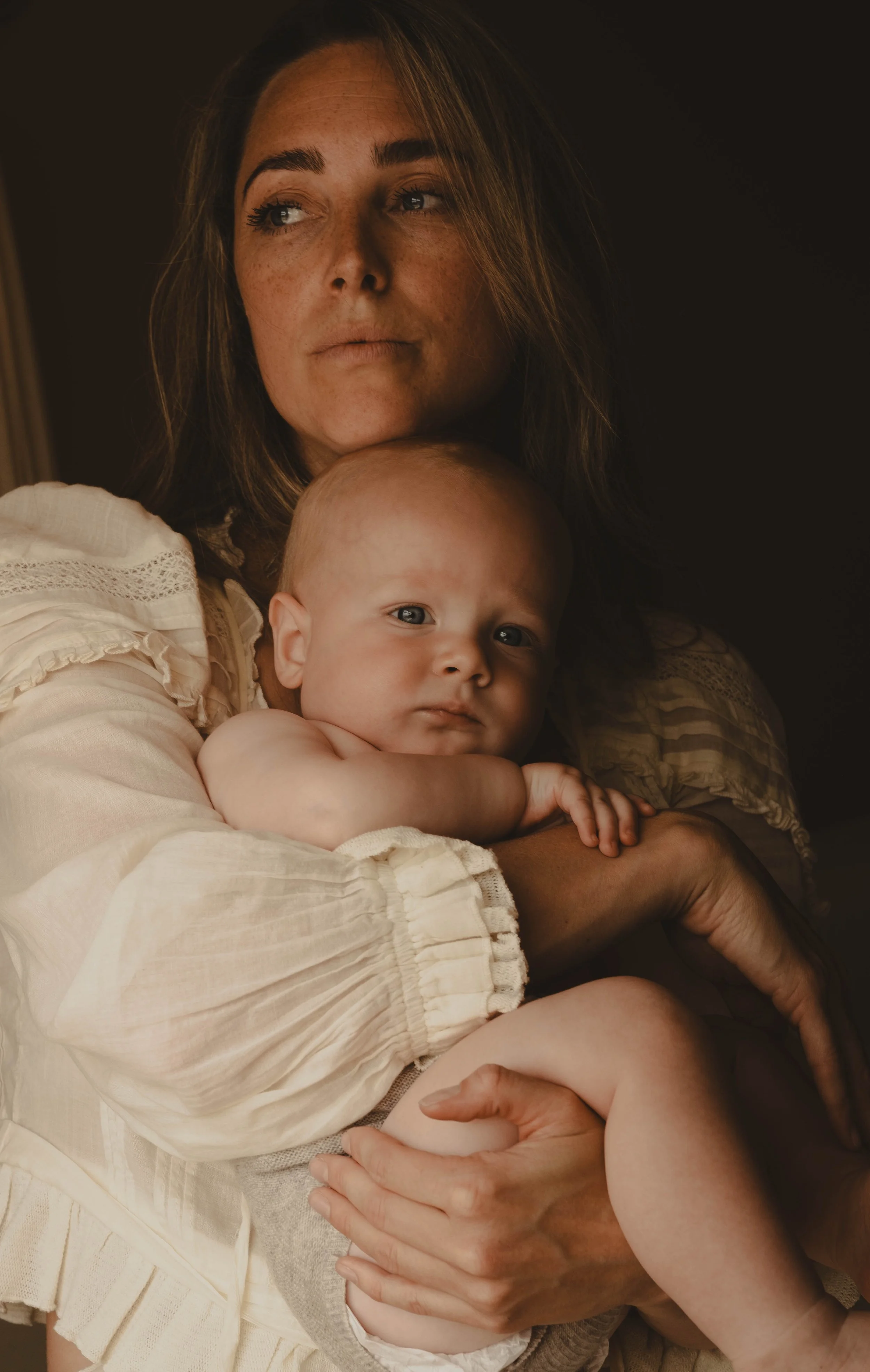 A woman holding a baby close to her chest, both looking at the camera. The woman has brown hair, and the baby has light skin and is resting on her arm with its head on her shoulder.