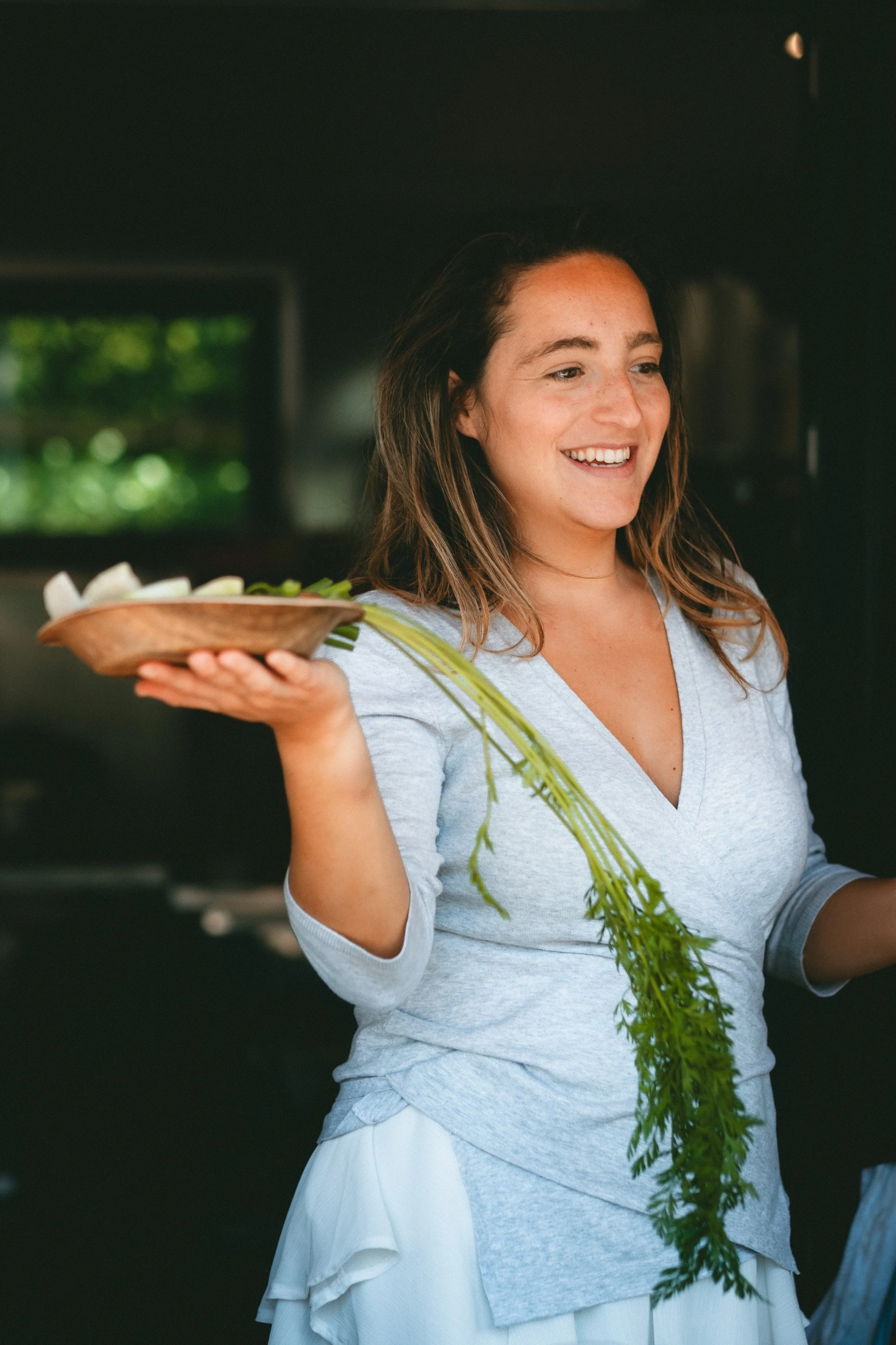 A woman with brown hair smiling and holding a wooden plate with flowers, standing indoors near a window with greenery outside.