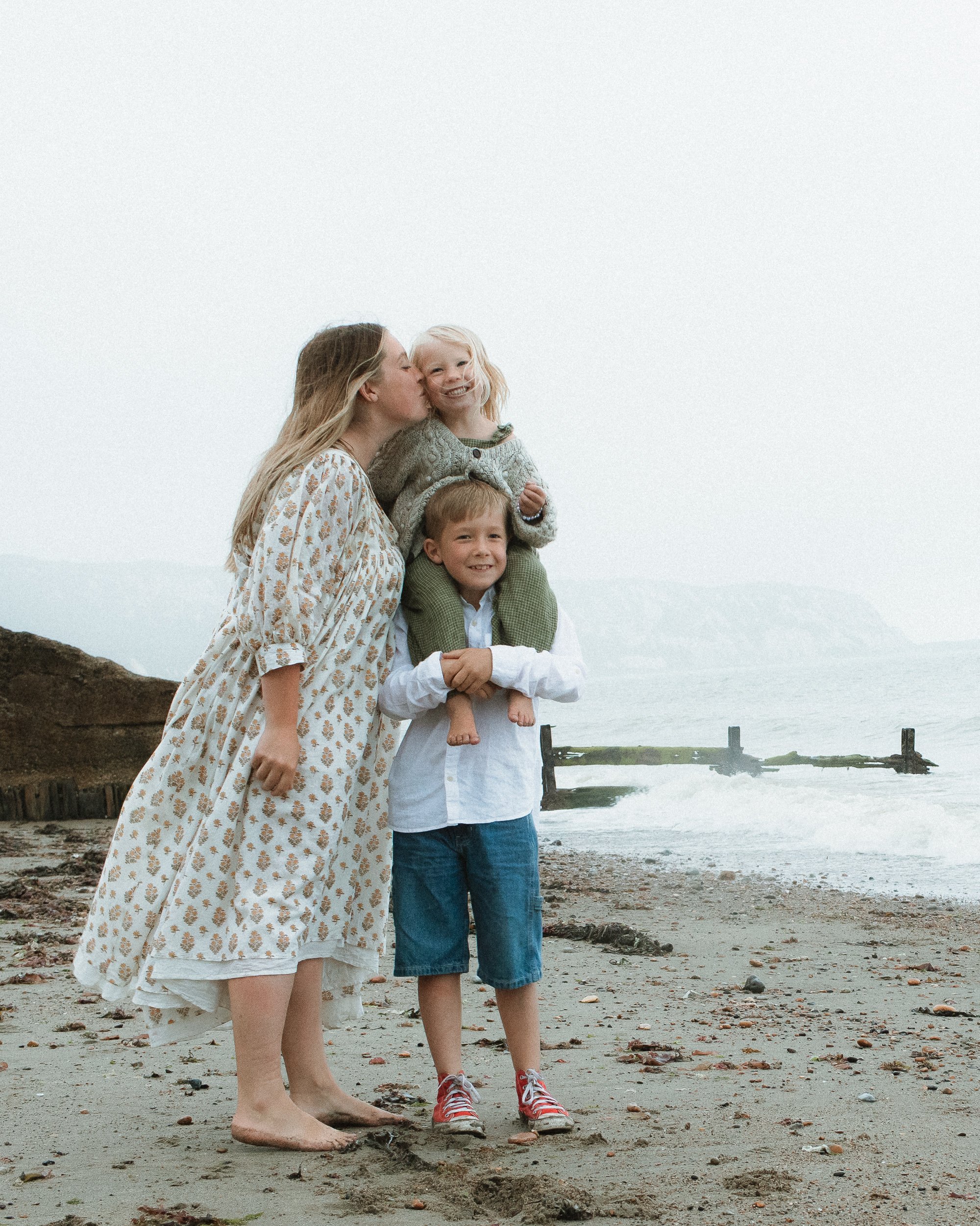A woman and two children standing on the beach, with the woman kissing a young girl on the cheek and the boy smiling, with a foggy ocean background.