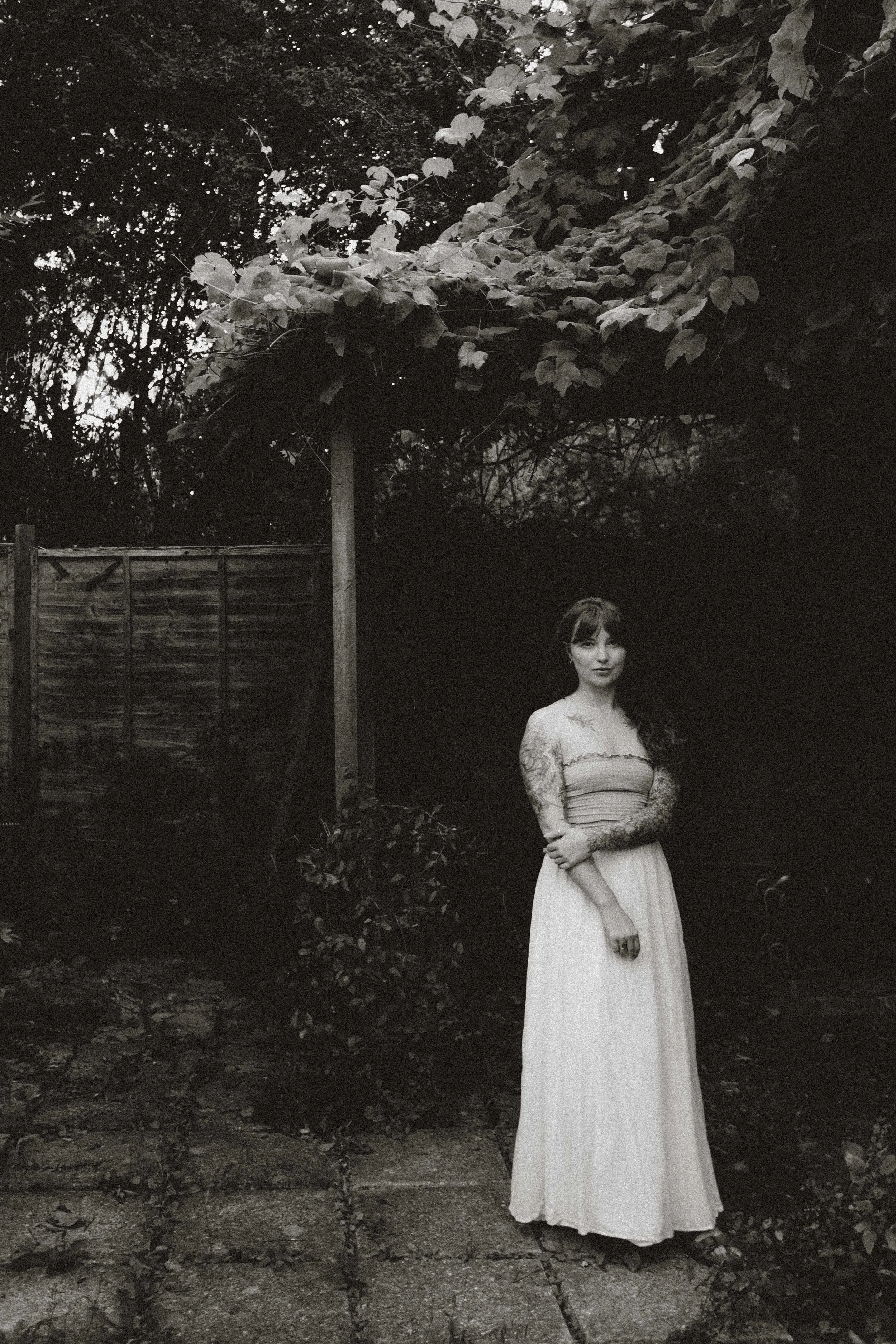 A woman with long dark hair and tattoos on her arms, wearing a strapless top and a long skirt, standing outdoors on a stone-paved path, with a wooden fence and trees in the background.