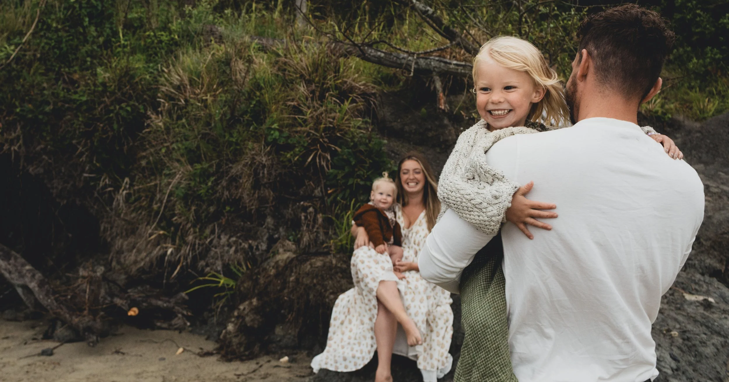 A man holding a smiling young girl in his arms at a beach, with a woman and a toddler sitting on rocks in the background, all smiling and enjoying the outdoor setting.