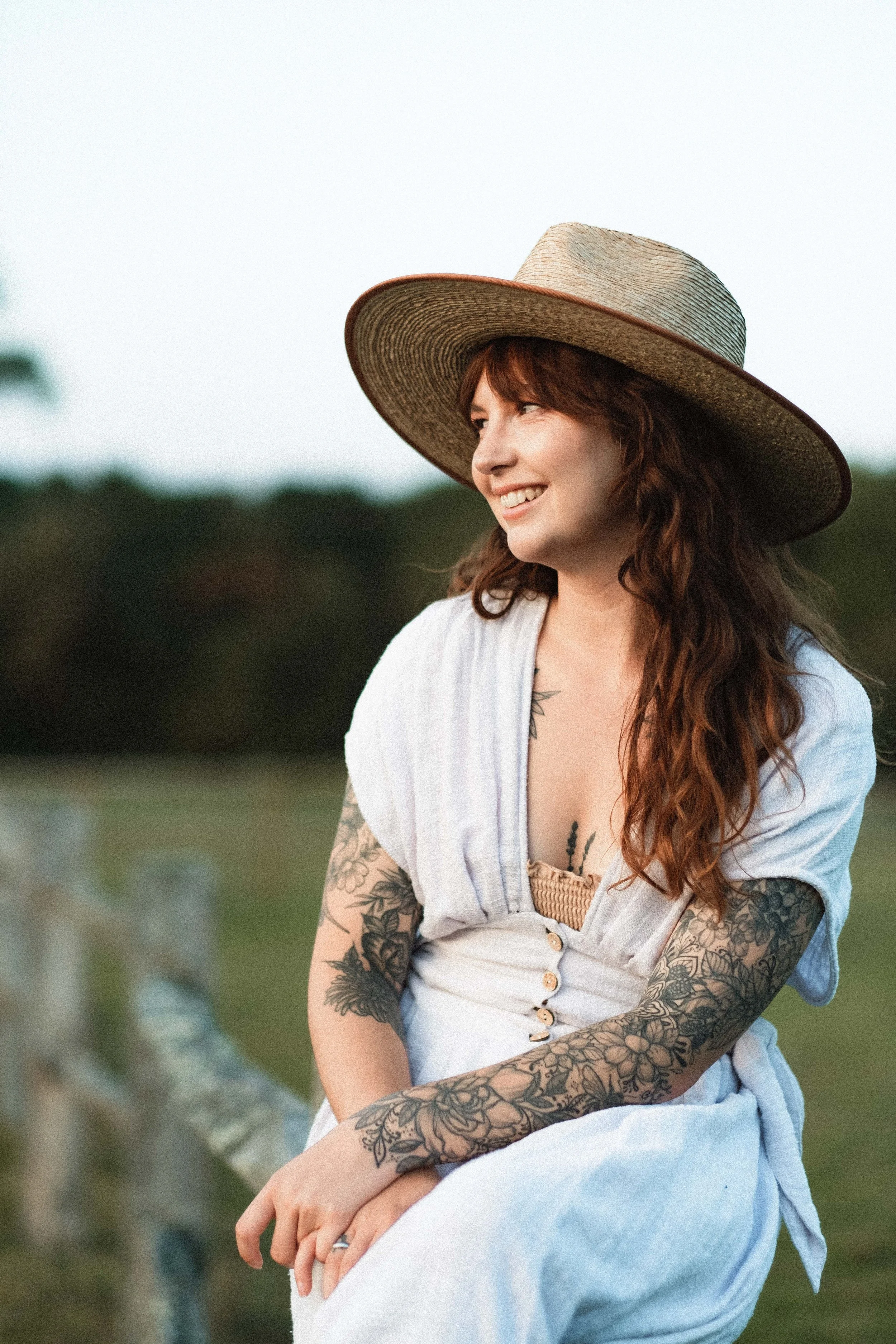 Smiling woman with tattoos on her arms and chest, wearing a large wide-brimmed straw hat and a white dress, sitting outdoors near a fence, with a blurred natural background.