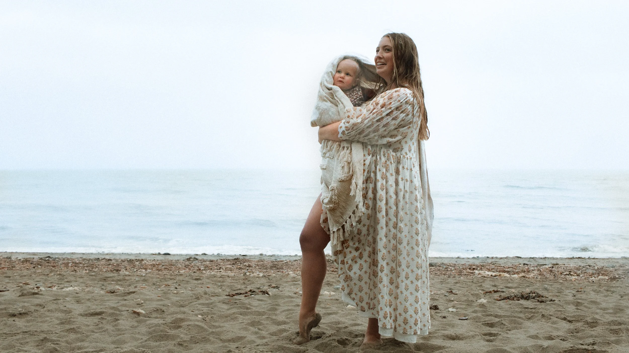 A woman holding a baby on a beach with the ocean and sky in the background.